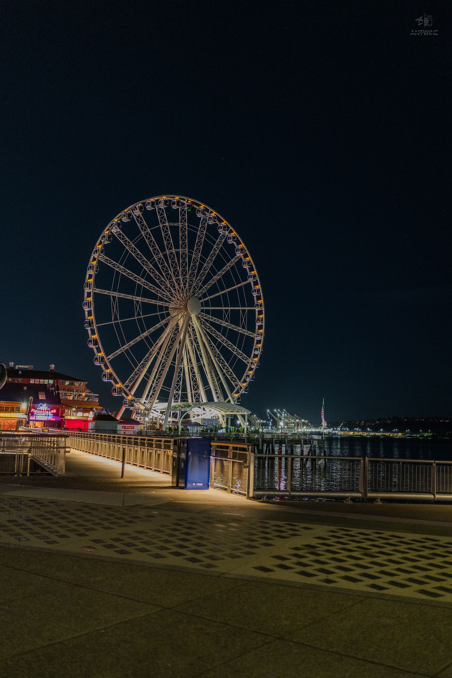 Illuminated Ferris wheel reflecting on waterfront at night with colorful lights against dark sky.