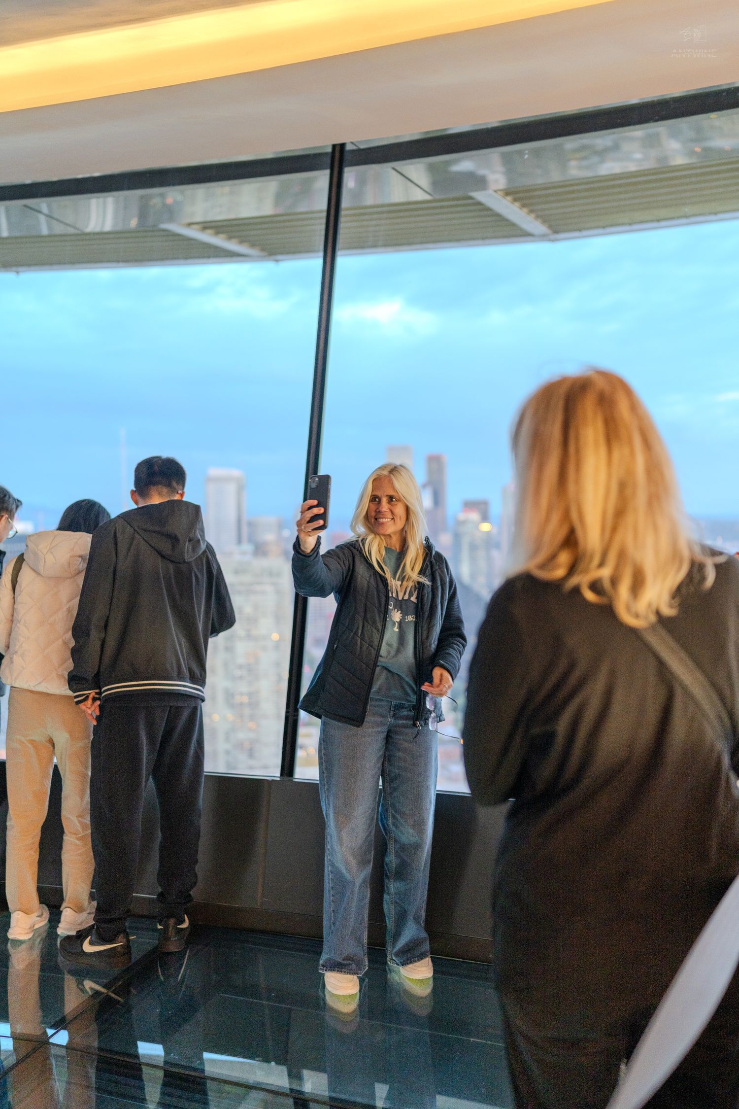 A blonde woman takes a selfie on a high-rise observation deck overlooking a city skyline at dusk.
