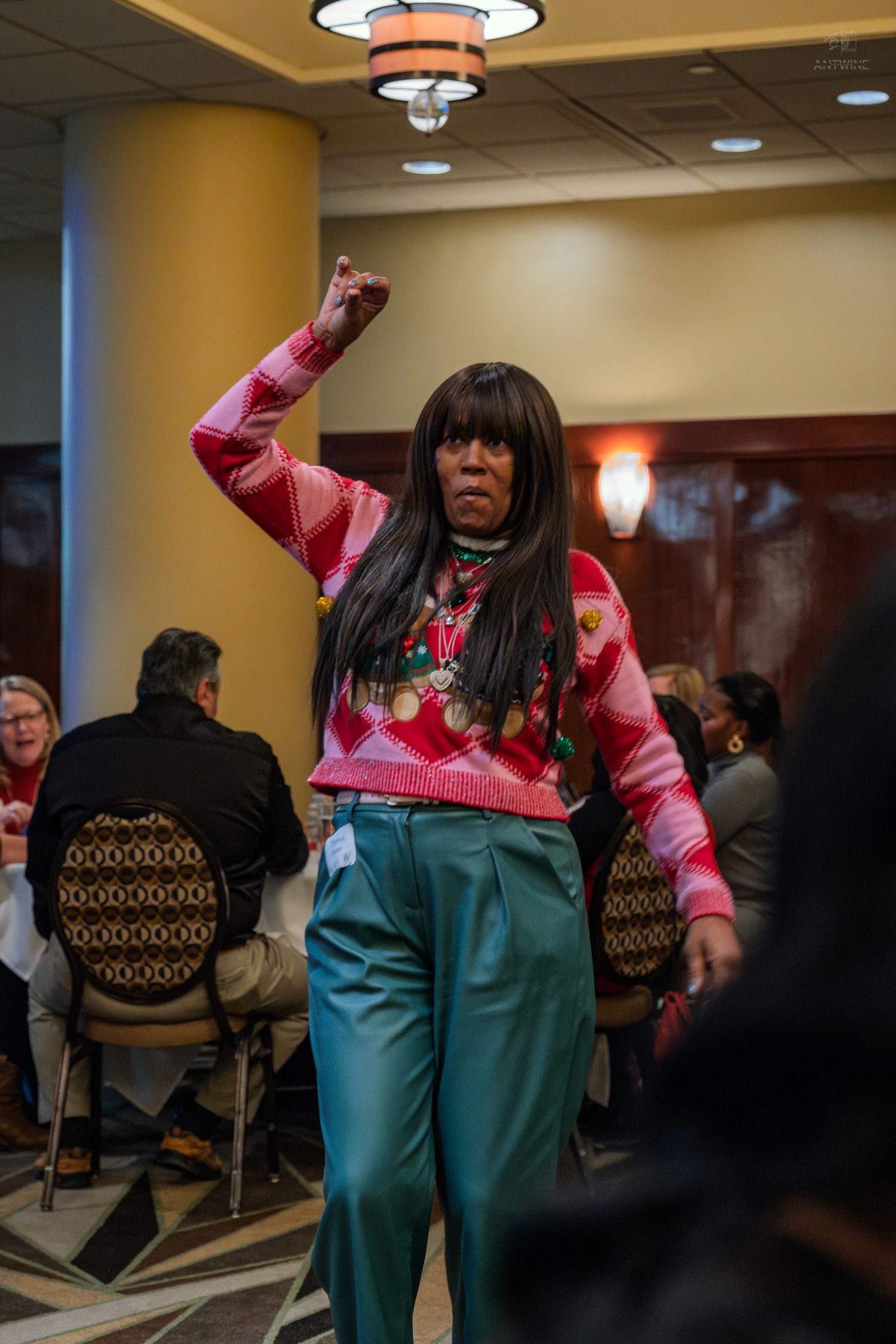 Woman in colorful pink sweater and teal pants raises fist confidently at an indoor banquet event.