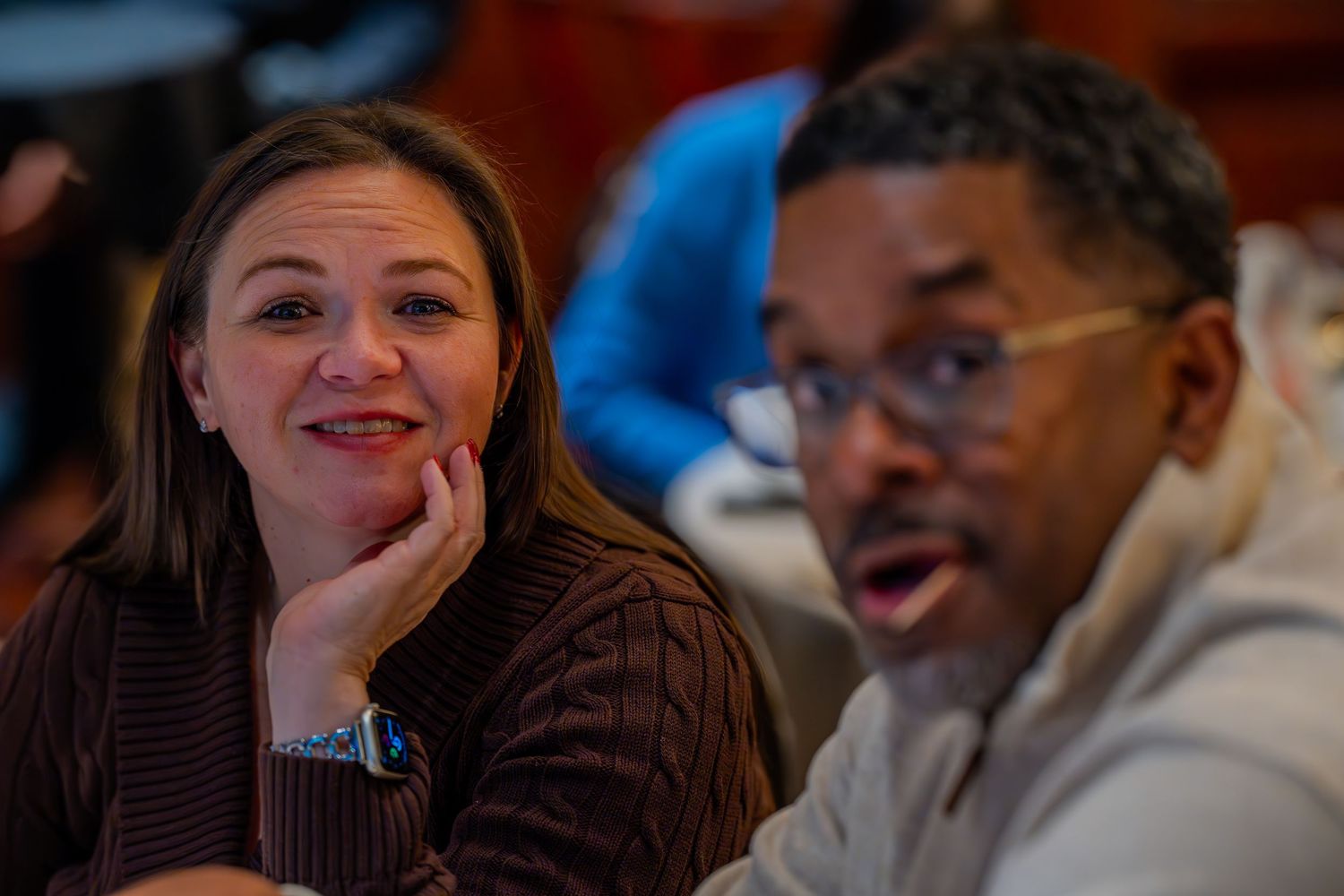 Two people engaged in conversation, a smiling woman and a man wearing glasses, seated together in a warmly lit room.
