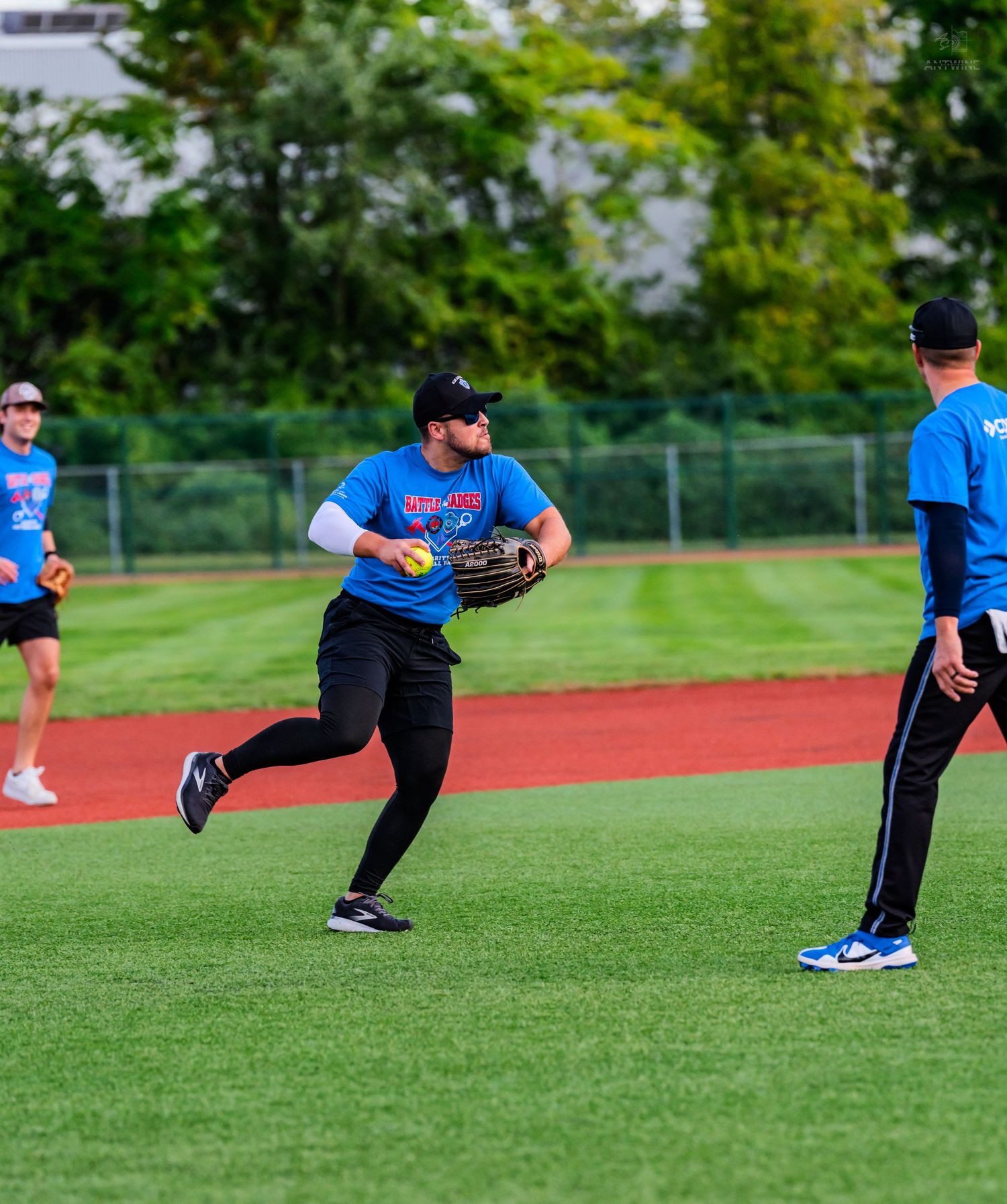 A bearded player in a blue shirt catches a ball while running on a green turf softball field.