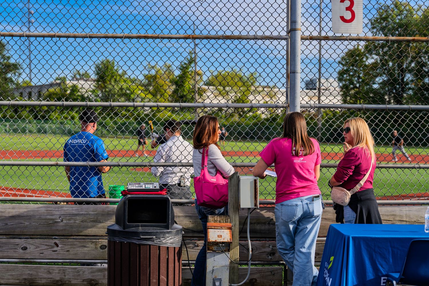 Spectators in pink shirts watch a baseball game through a chain-link fence at an outdoor sports field.