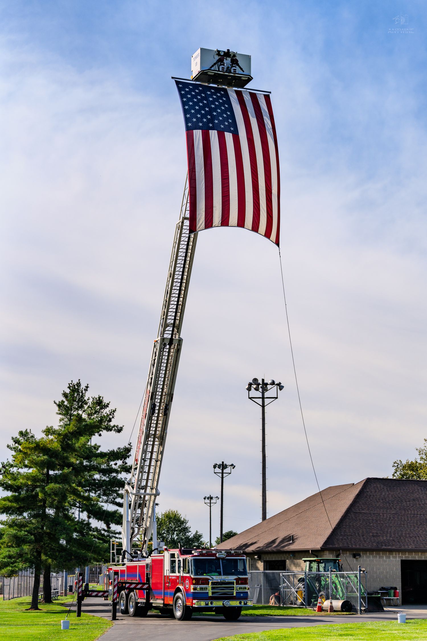 Fire truck ladder extended high displaying large American flag against blue sky near a building and green trees.
