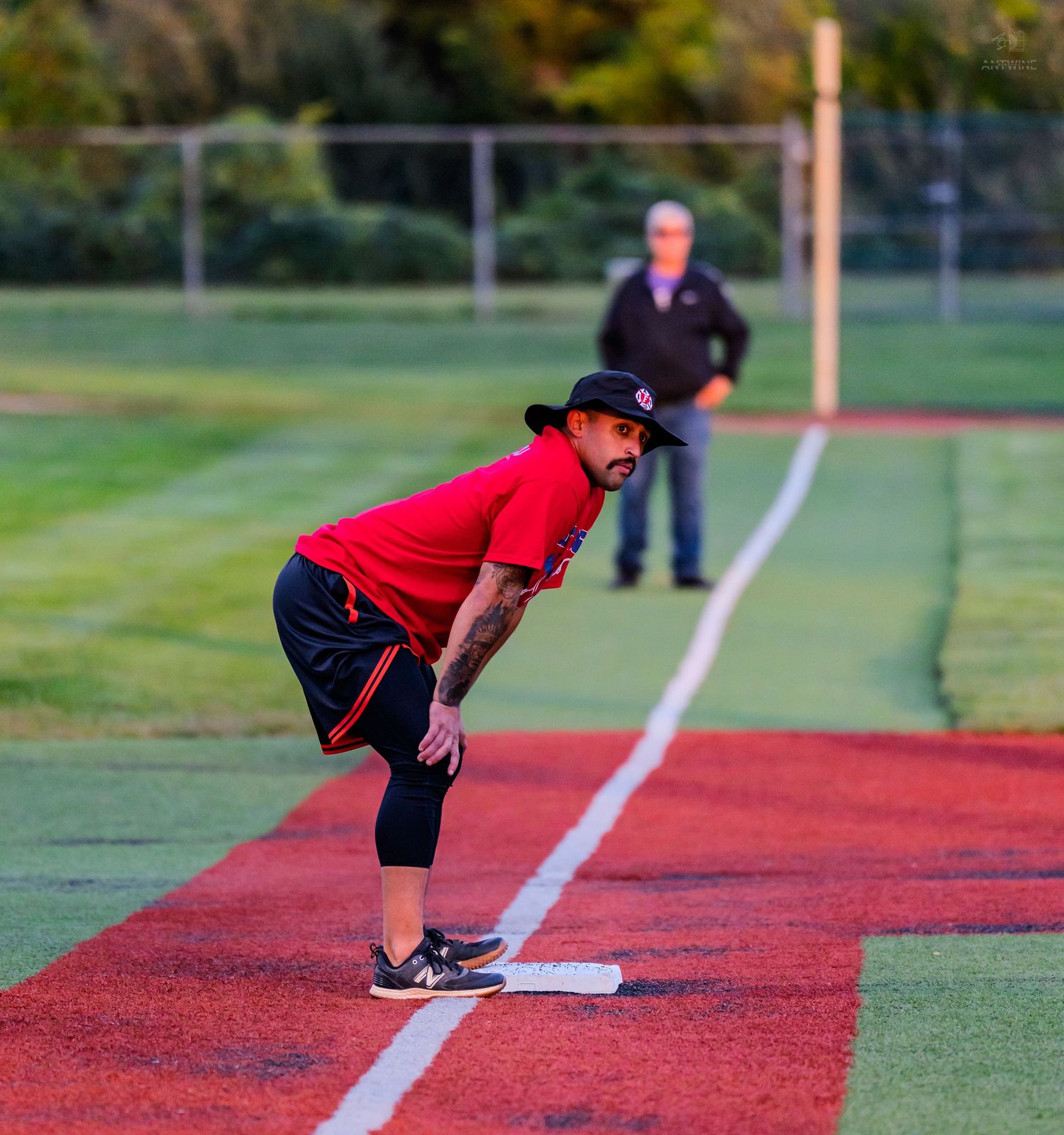 A softball player in red uniform crouches at base during a game, with an umpire watching in the background.
