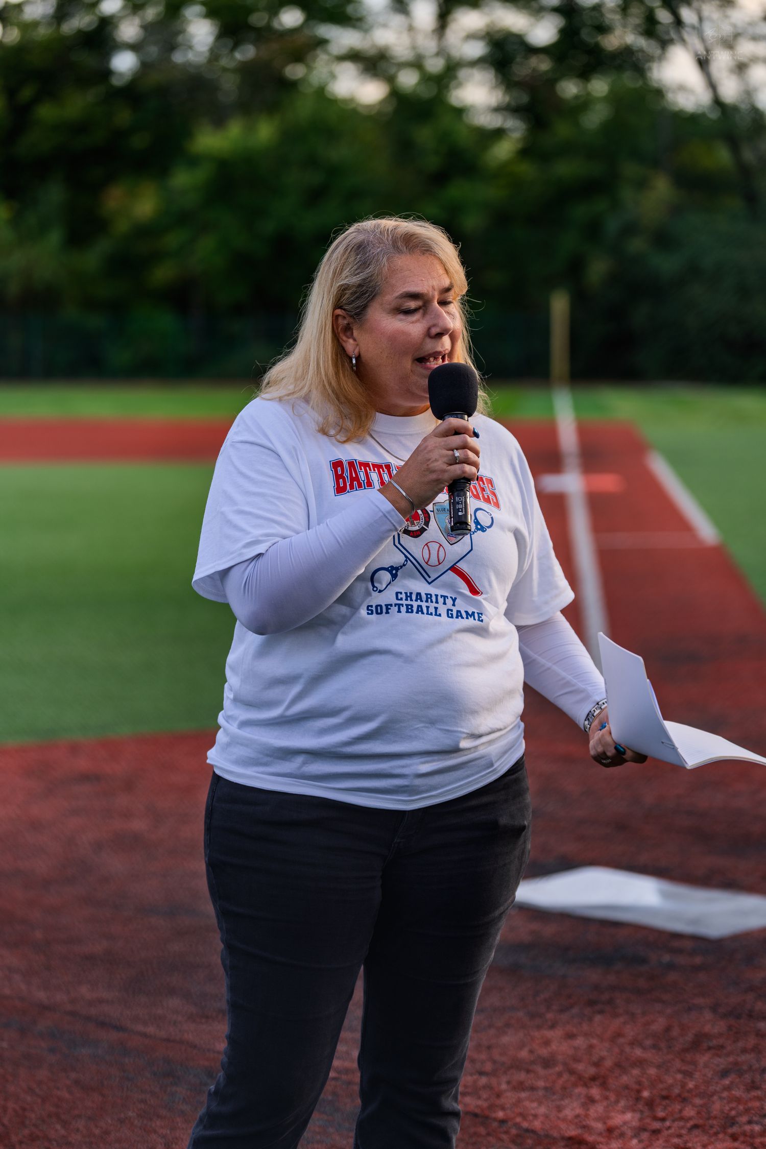 Woman speaking into microphone at charity softball game on baseball field wearing event t-shirt.