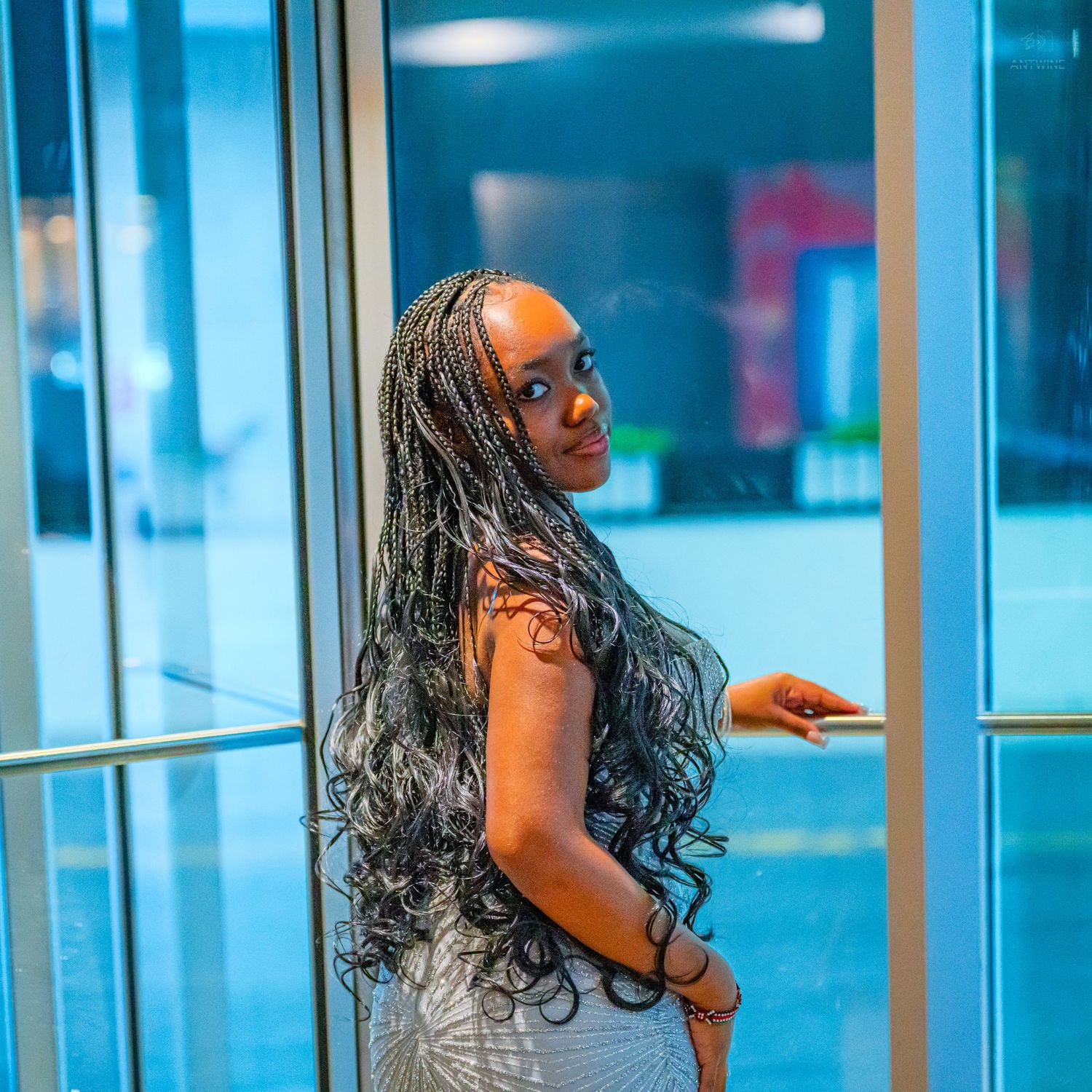 Young woman with long braided hair poses elegantly in a glass doorway wearing a white dress.