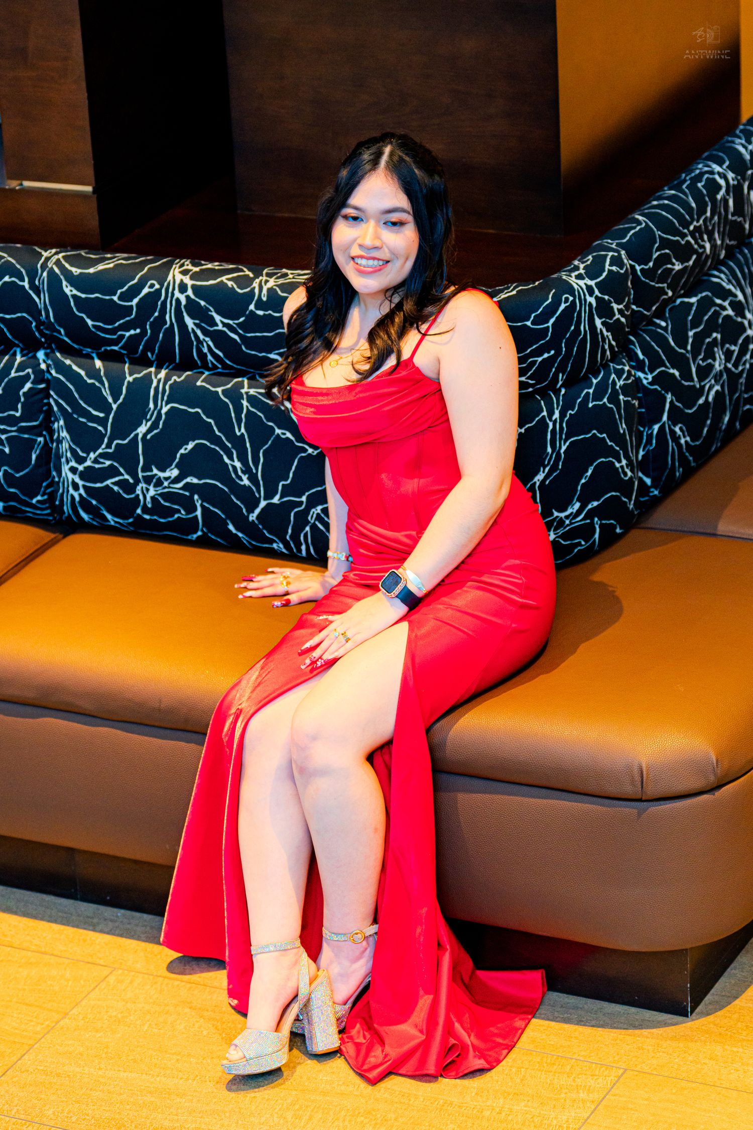 A smiling young woman in a red slit gown and heels sits elegantly on a brown leather sofa.