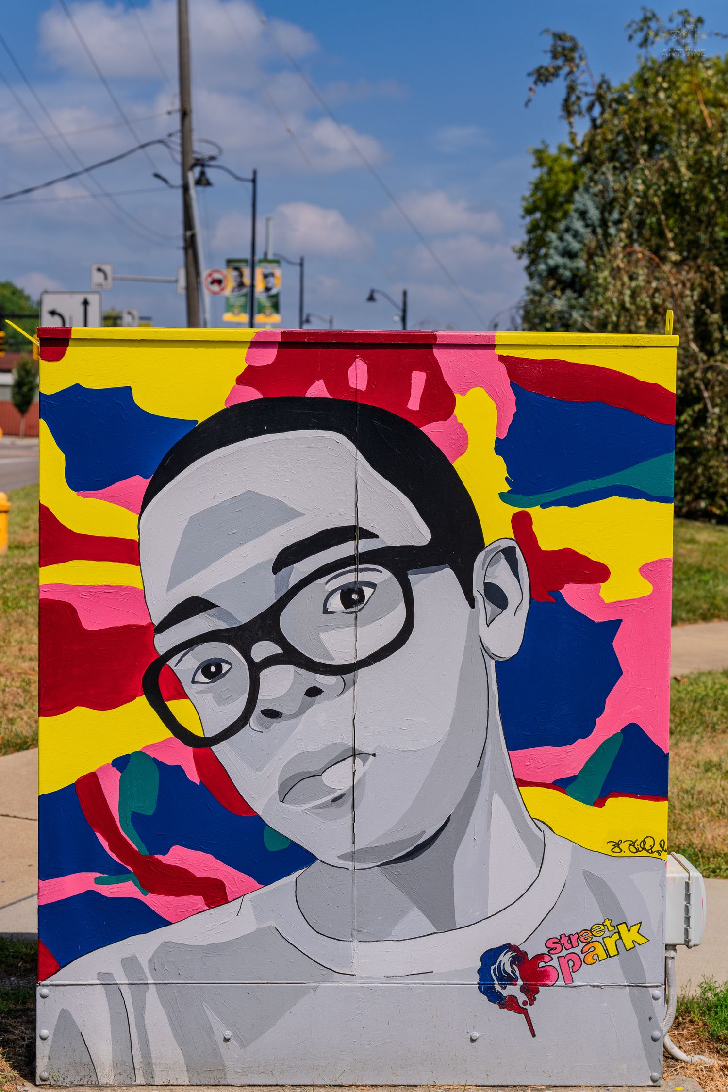 Colorful street art mural on utility box featuring a young person wearing glasses with vibrant yellow, pink, and blue background.