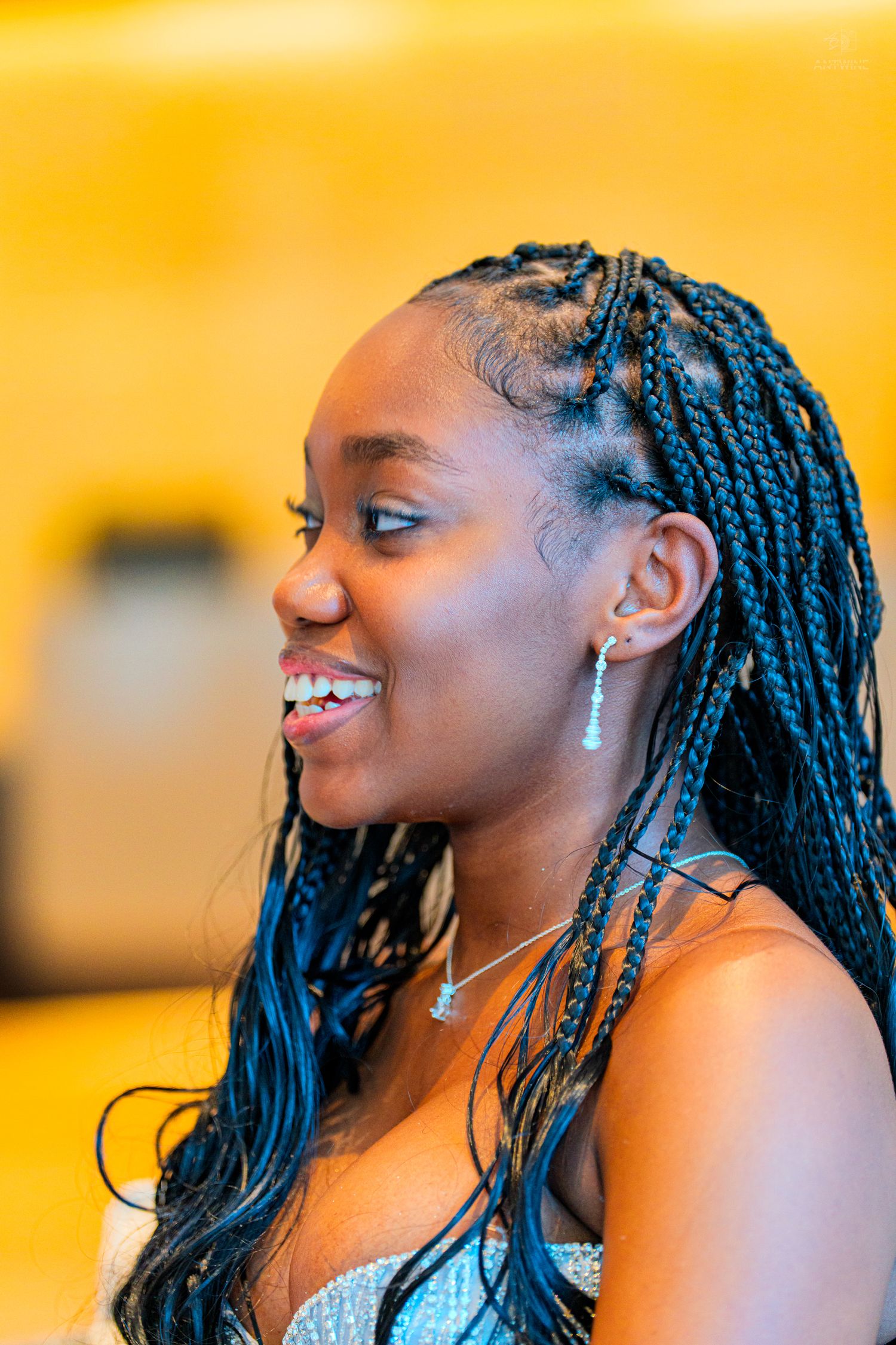 Young woman with long box braids smiling joyfully, wearing elegant drop earrings and necklace against yellow background.
