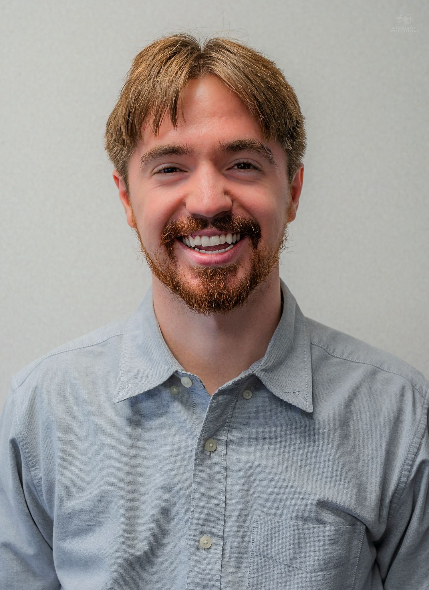 Smiling young man with reddish-brown hair and beard wearing a light blue button-up shirt against a gray background.