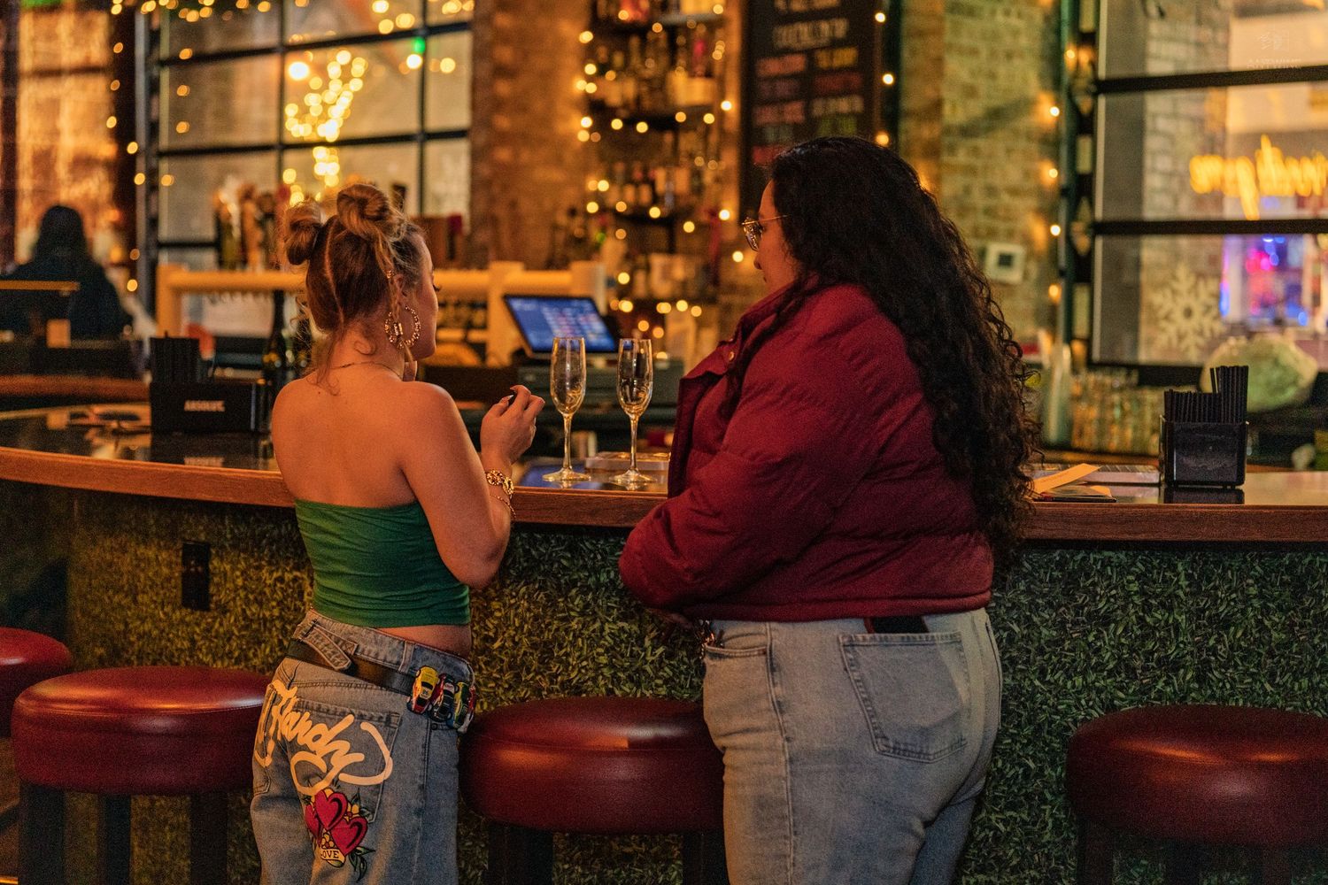 Two women chatting at a stylish bar counter with warm string lights in the background.