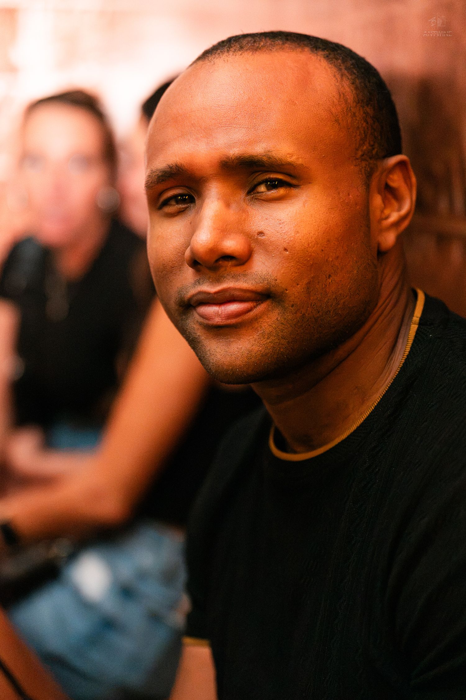 A man in a black t-shirt smiles at the camera in a warmly lit venue.