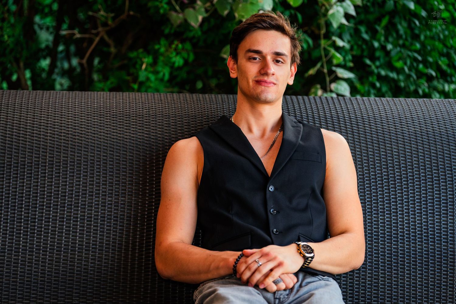 Young man in black sleeveless shirt sitting relaxed on wicker bench surrounded by lush green foliage.