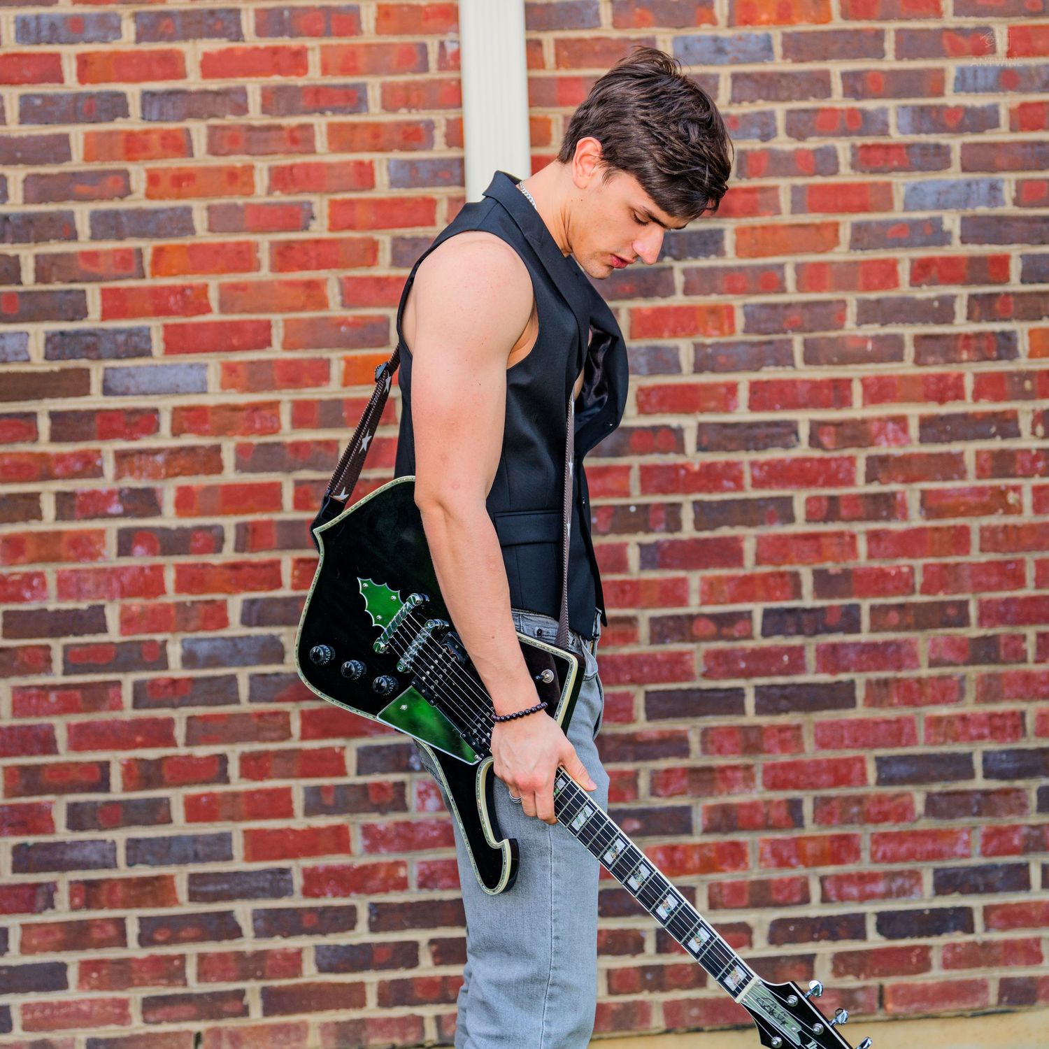 Young male guitarist in sleeveless vest holding black electric guitar against brick wall.