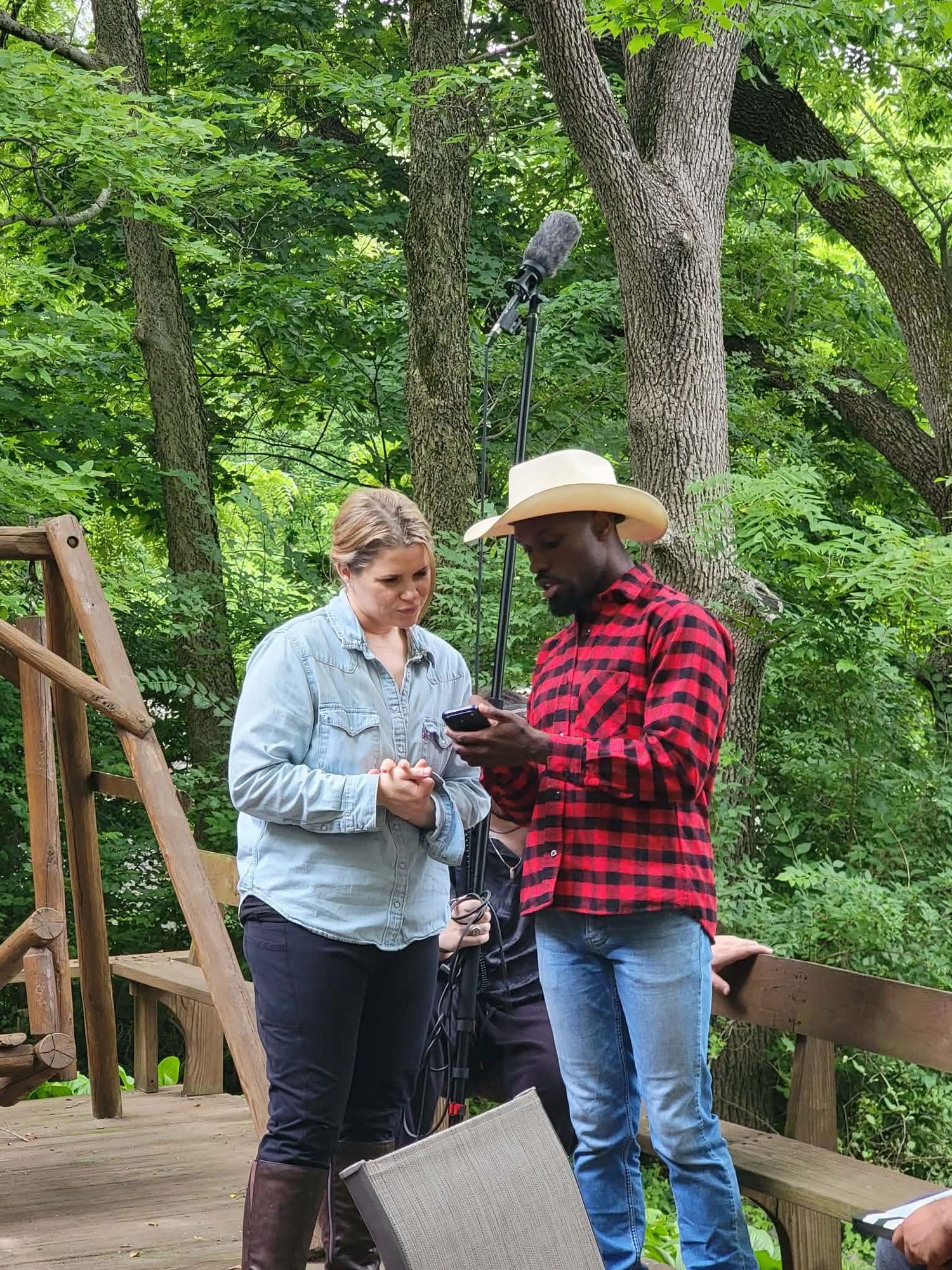 Woman in denim and man in plaid cowboy hat review phone content on a wooden deck in the woods.