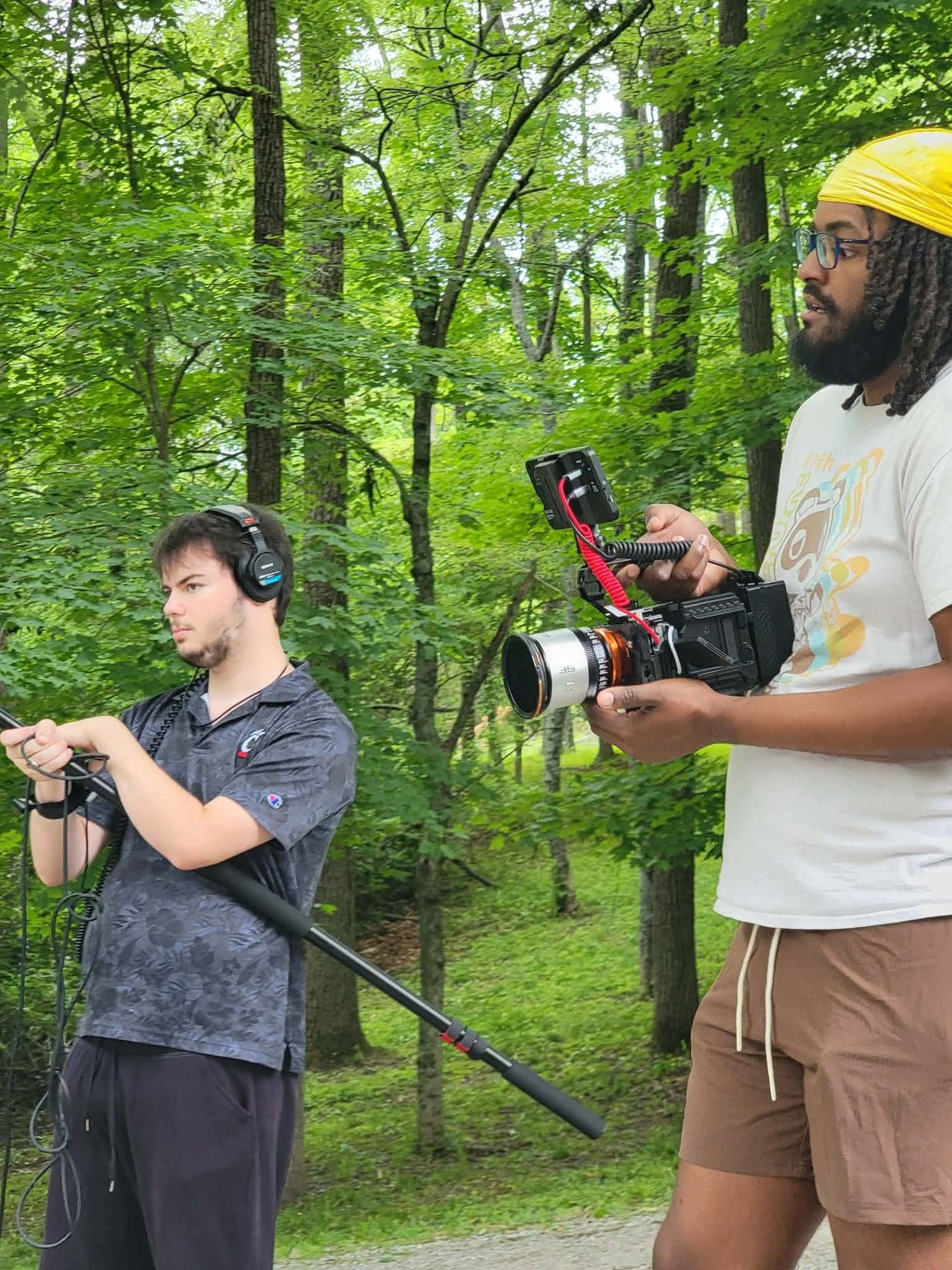Two filmmakers work outdoors in a forest, one holding a boom mic and the other operating a cinema camera.