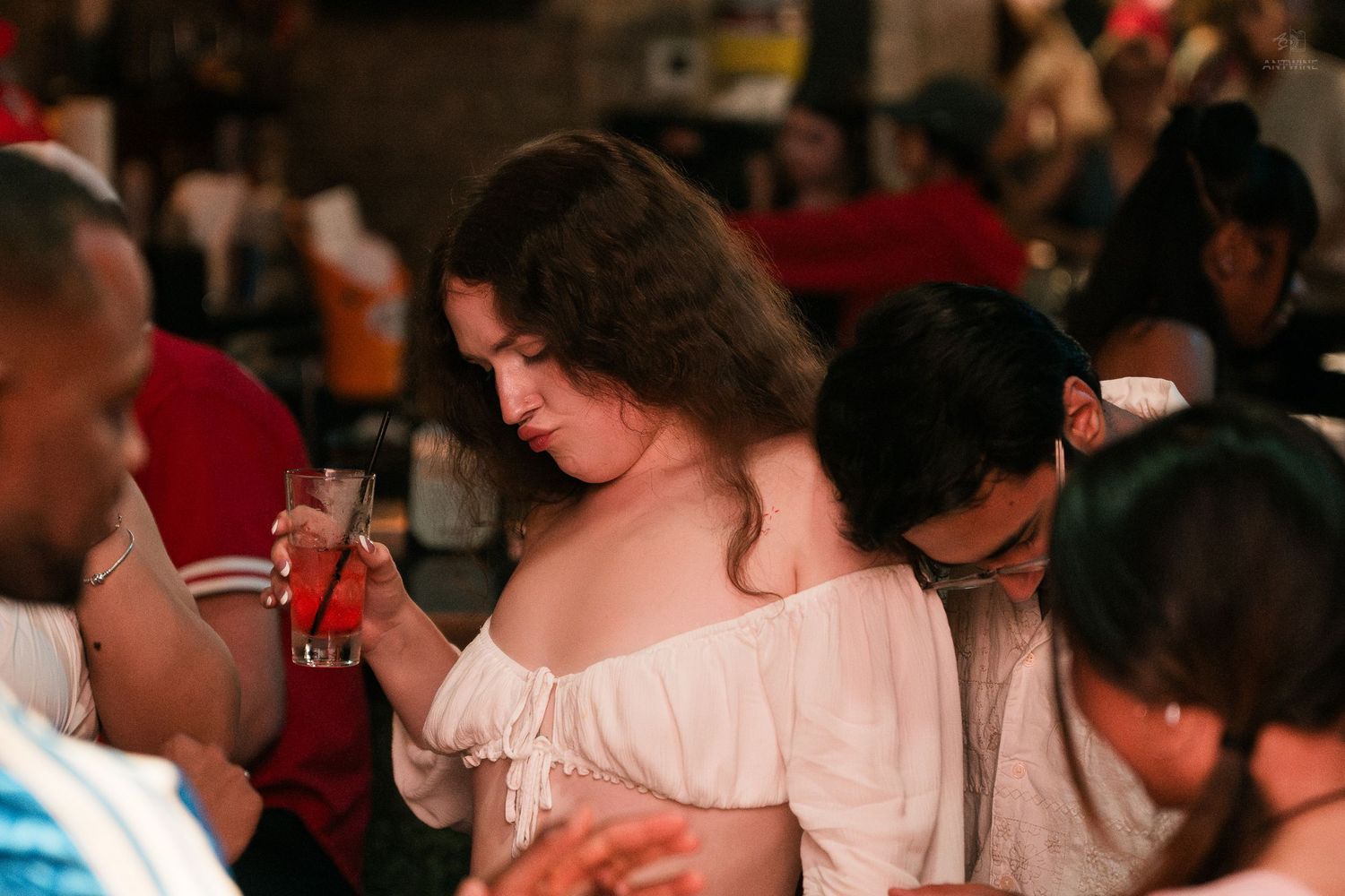 Woman in white off-shoulder top holding a cocktail at a crowded bar gathering.