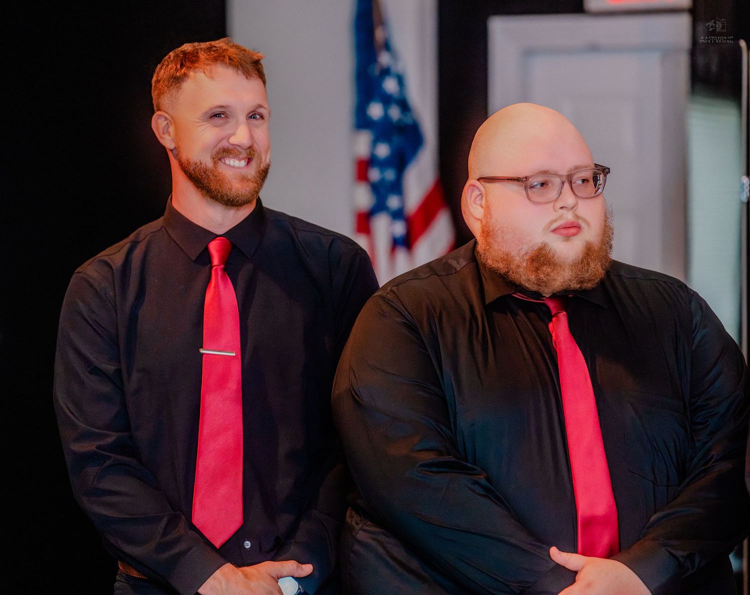 Two groomsmen in black shirts and red ties smile during a wedding reception ceremony.