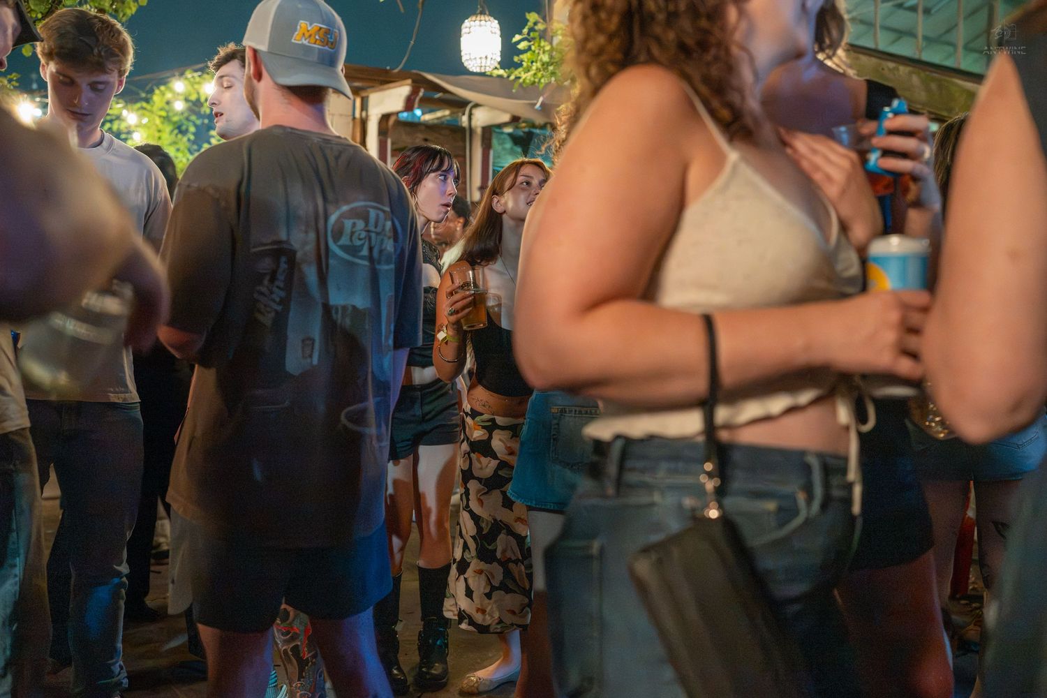 Crowd of people socializing outdoors at a nighttime festival event, holding drinks in a casual summer setting.