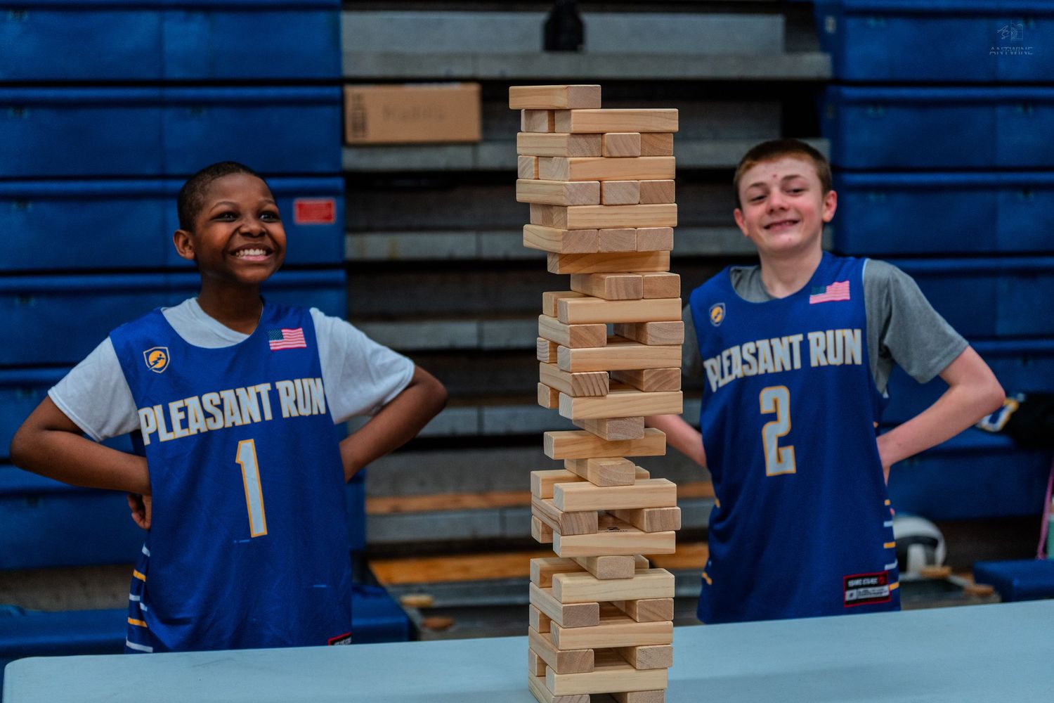 Two Pleasant Run basketball players smile beside a tall Jenga tower in a school gymnasium.