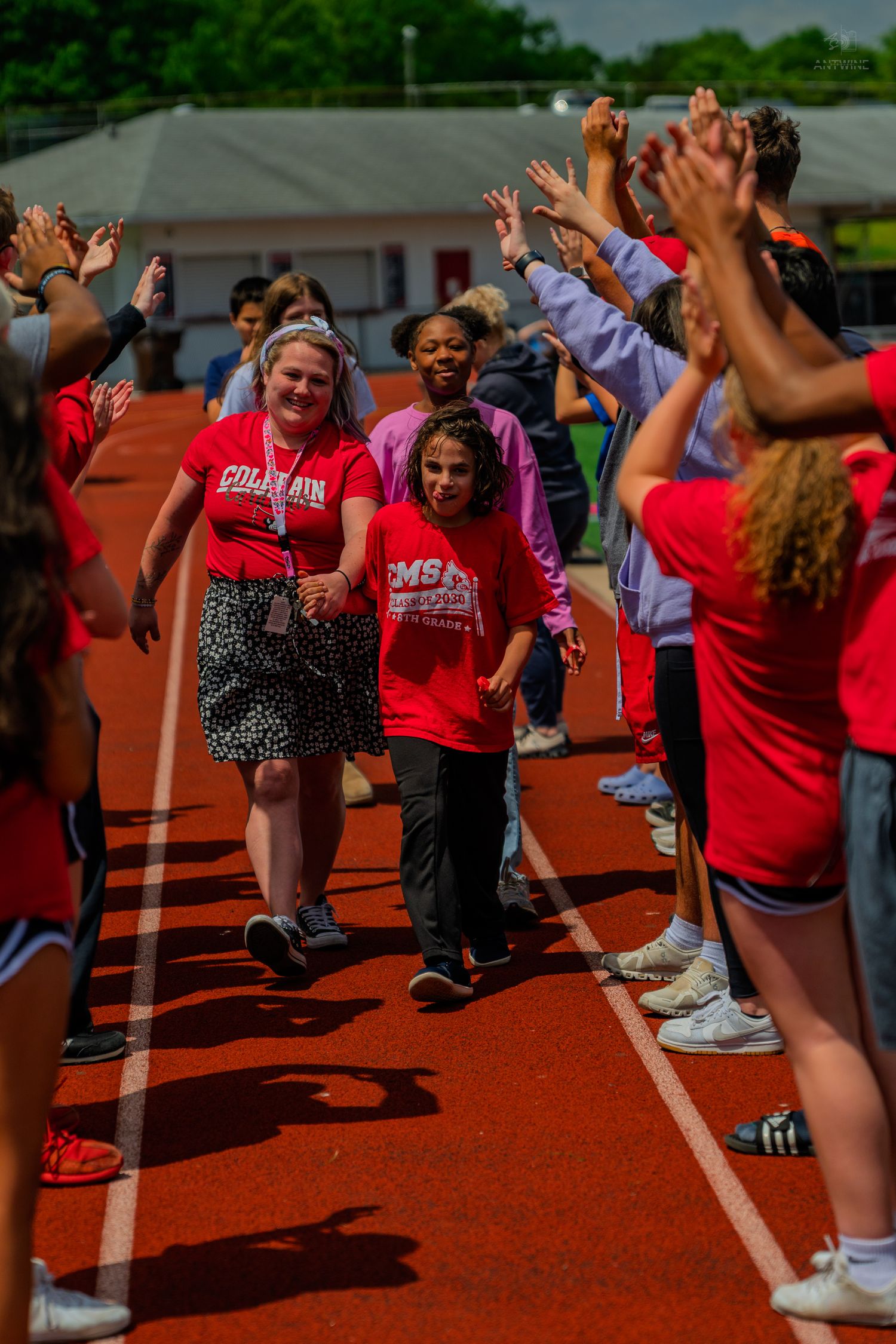 Students in red shirts walk through a celebratory high-five tunnel on a red track during a school sports event.