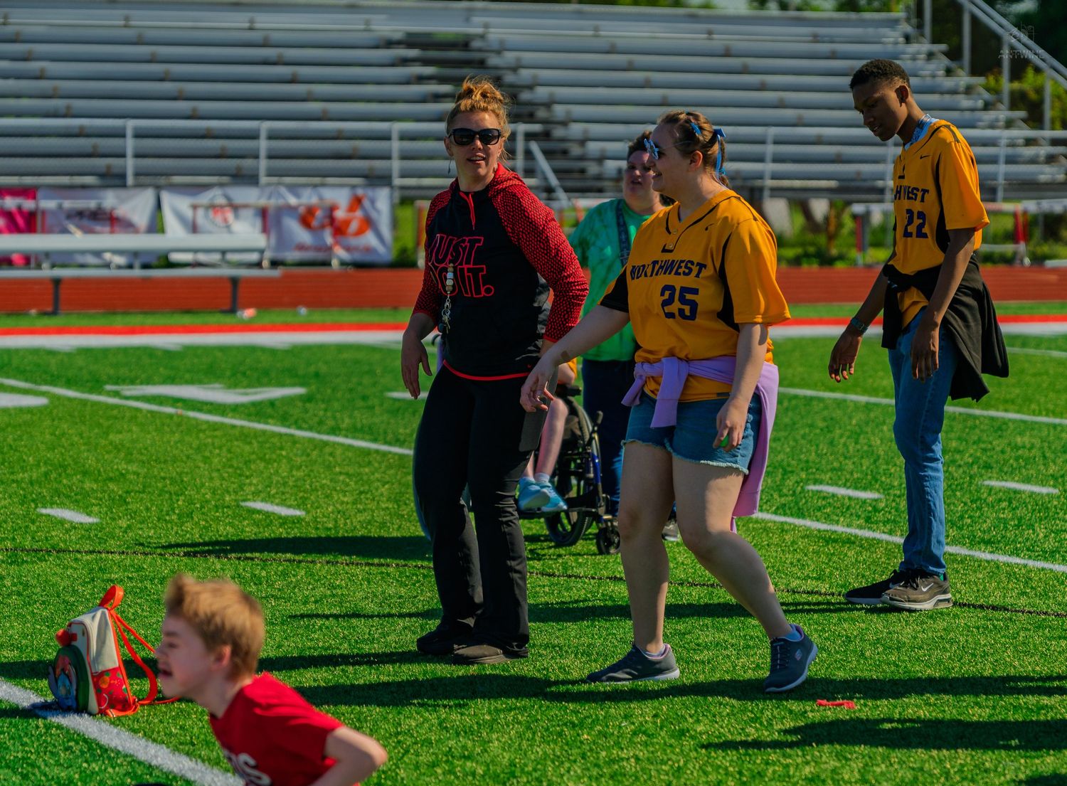 People in athletic wear on a green sports field, including a child crawling and adults wearing numbered jerseys.