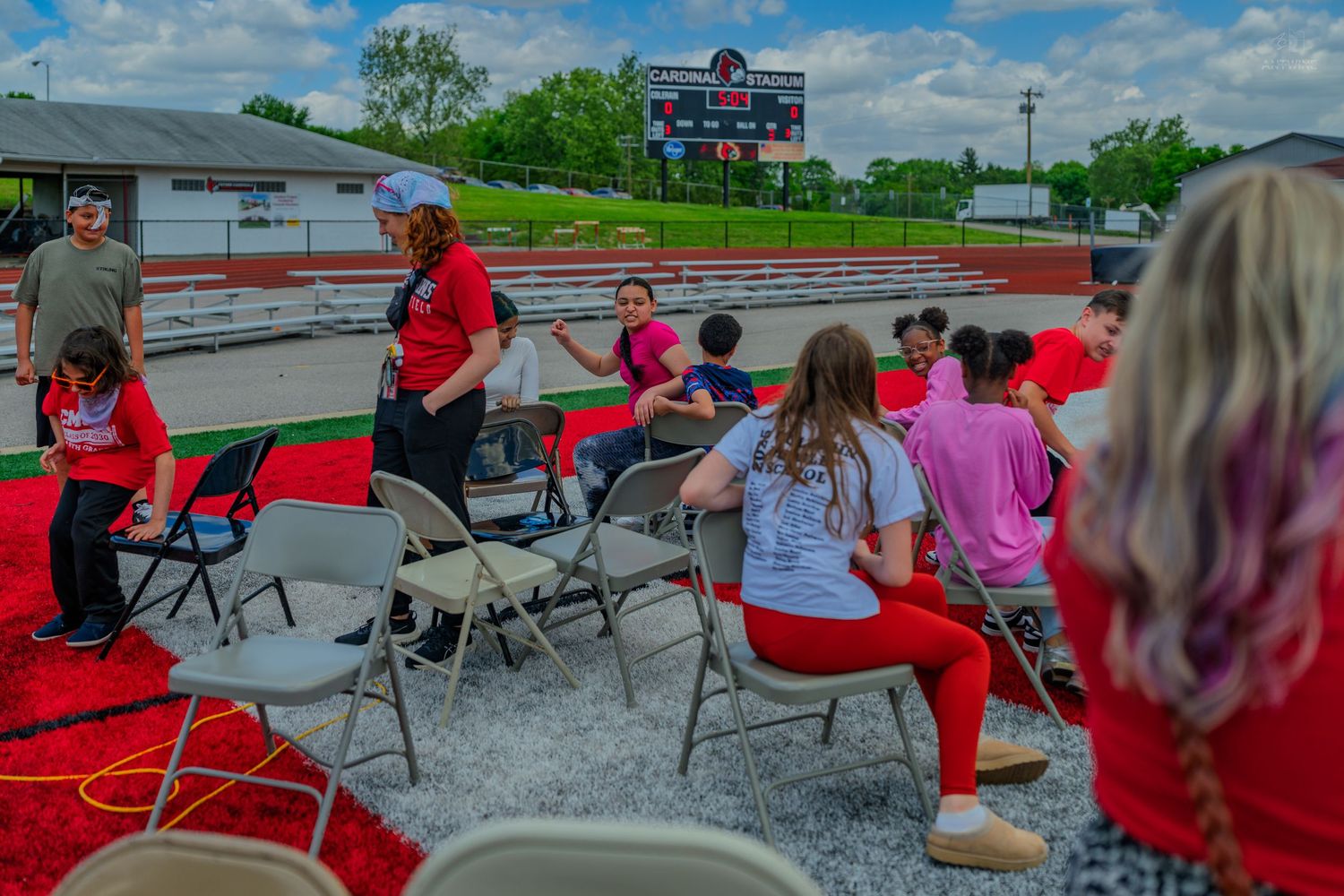 Group of people in red shirts socializing on outdoor track with folding chairs and scoreboard backdrop.