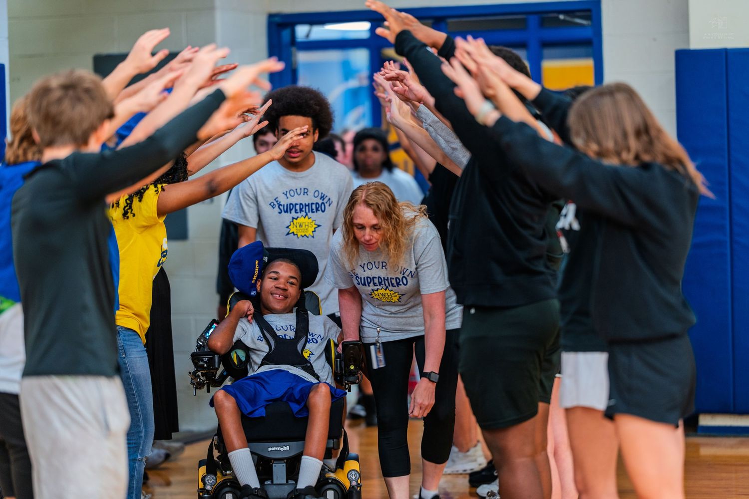 Students form a celebratory tunnel for a peer in a wheelchair during a Special Olympics event in a school gym.