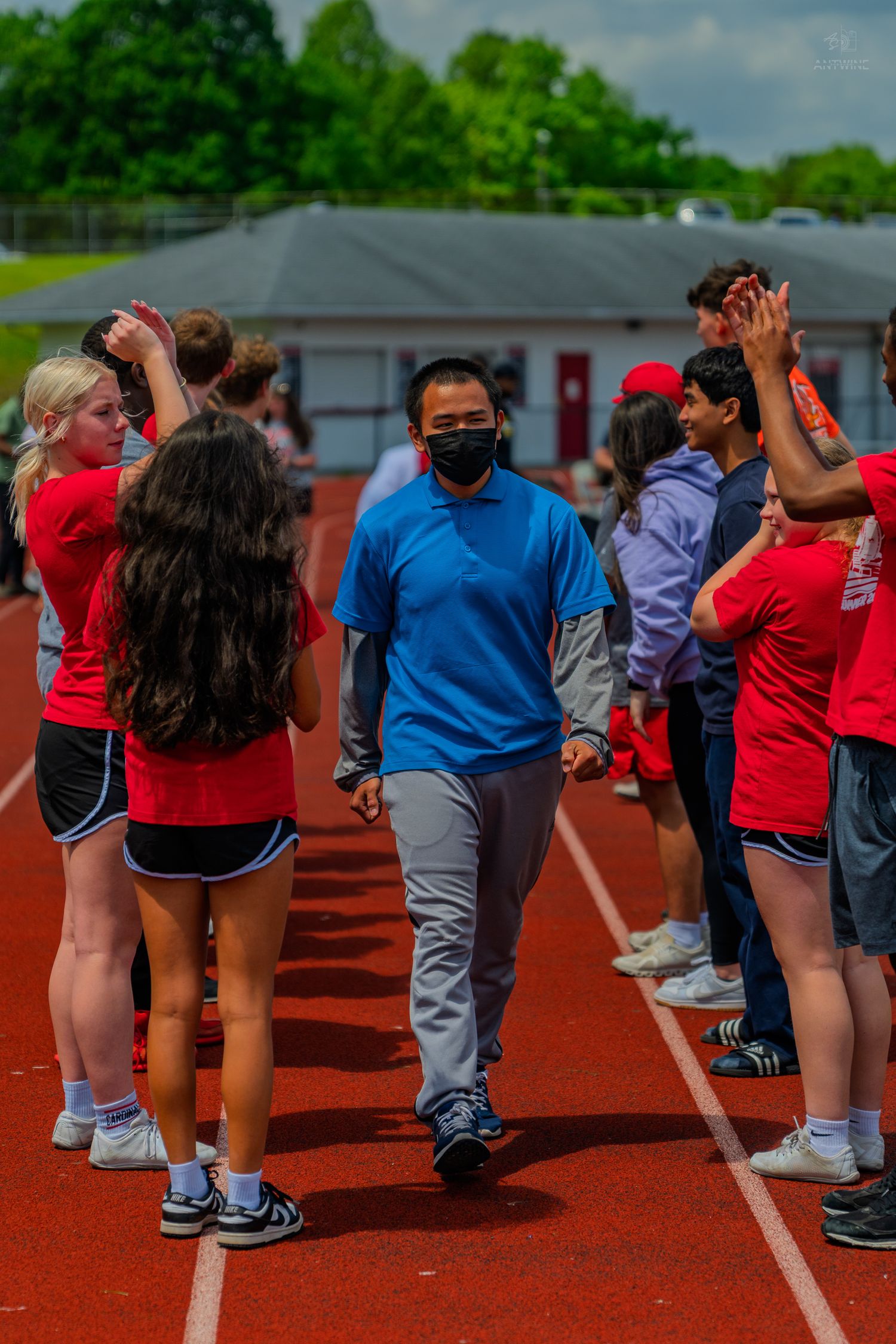 Man in blue shirt and mask walks through a high-five line of students in red on a school track.
