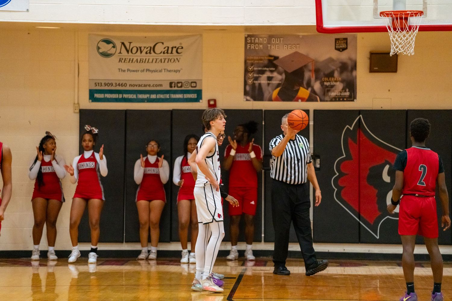 Basketball player stands at free throw line with referee and cheerleaders visible near Cardinals gym wall.