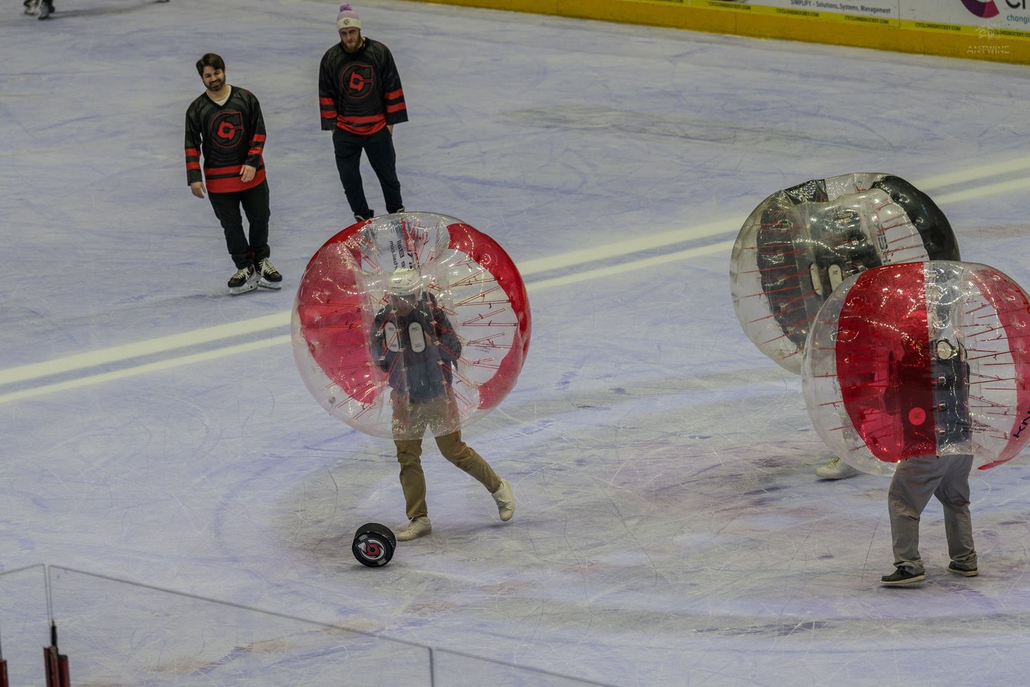 Hockey players watch as participants in transparent zorb balls play bubble soccer on an ice rink.