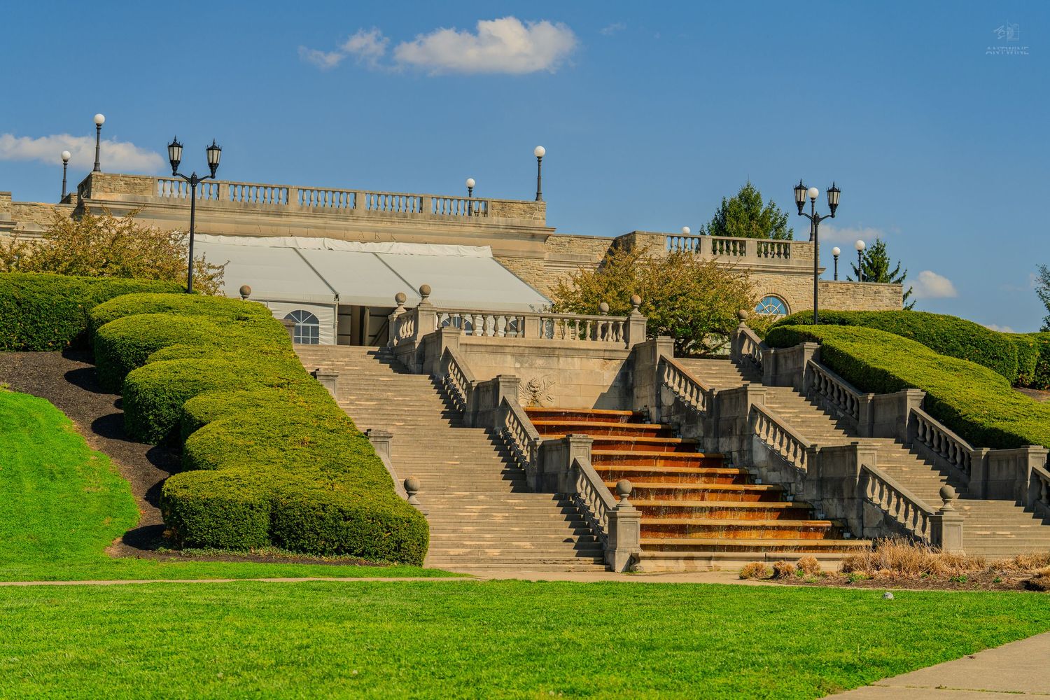 Grand stone staircase with ornate railings leading to a classical terrace building surrounded by manicured hedges and green lawn.