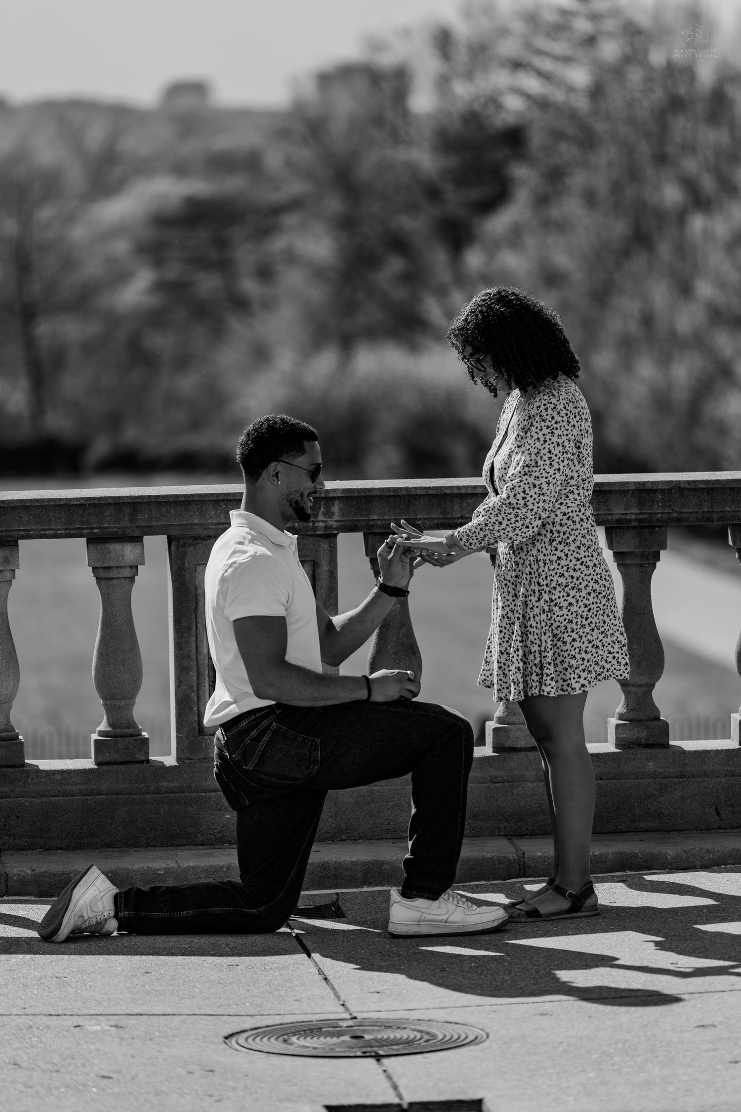 Black and white romantic marriage proposal with man on one knee holding womans hand outdoors.