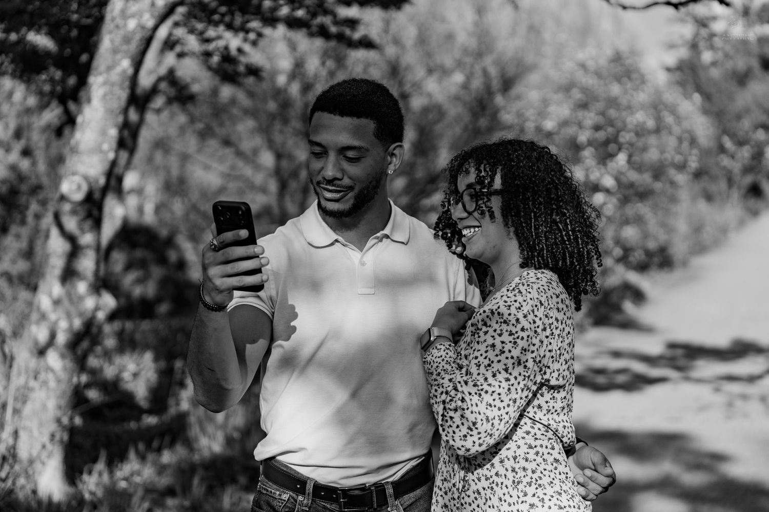 Black and white photo of smiling couple viewing smartphone together on a tree-lined outdoor path.
