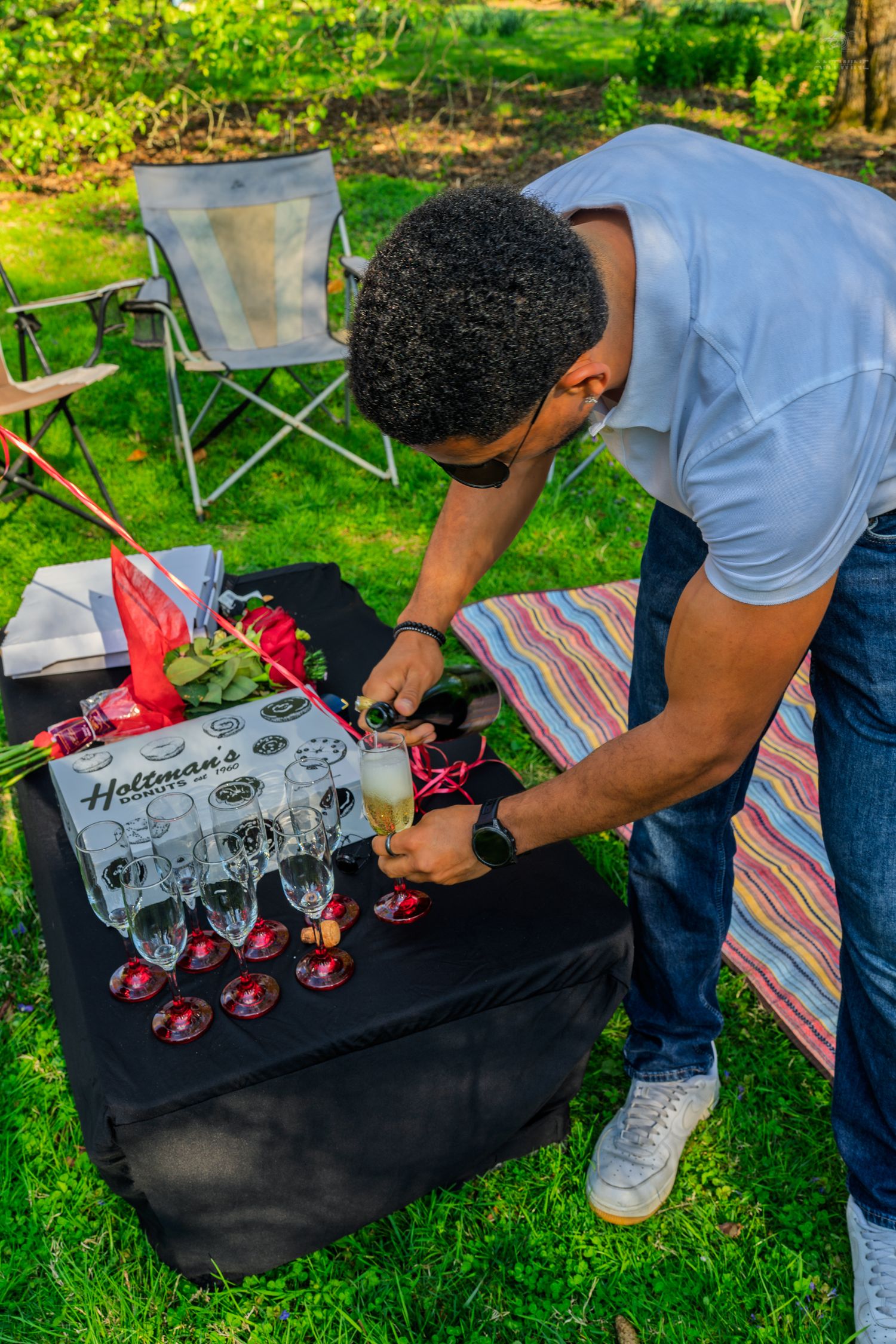 Man pouring sparkling wine into glasses at Holtman's branded outdoor picnic table setup in garden.
