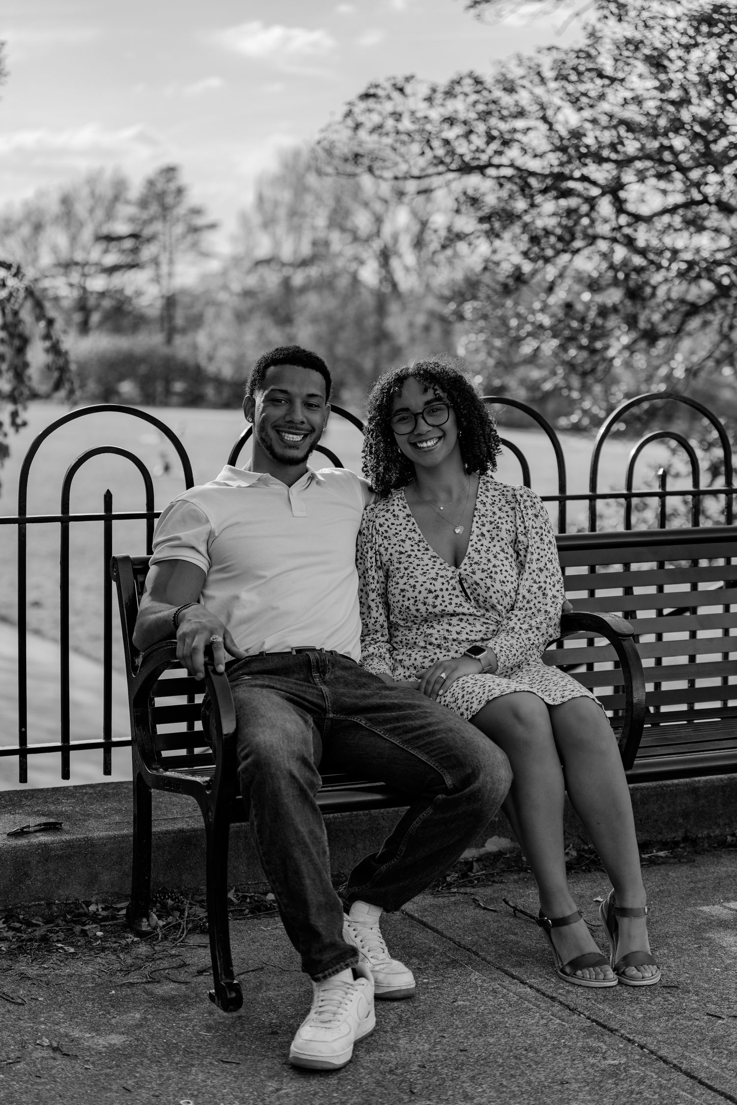 Black and white photo of a laughing couple sitting on a park bench with iron railing behind them.