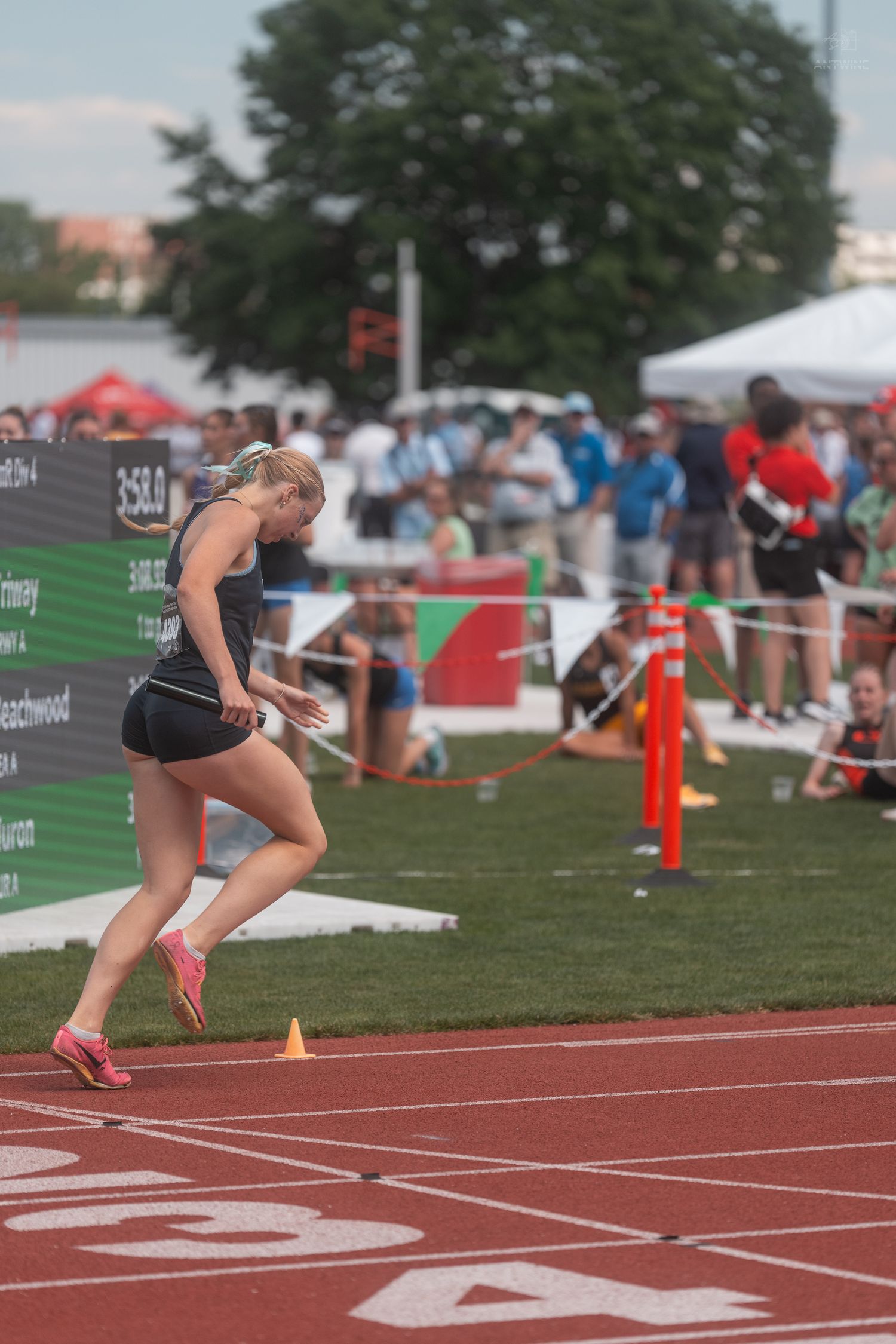Blonde female sprinter in black uniform crosses finish line at outdoor state track championship event.