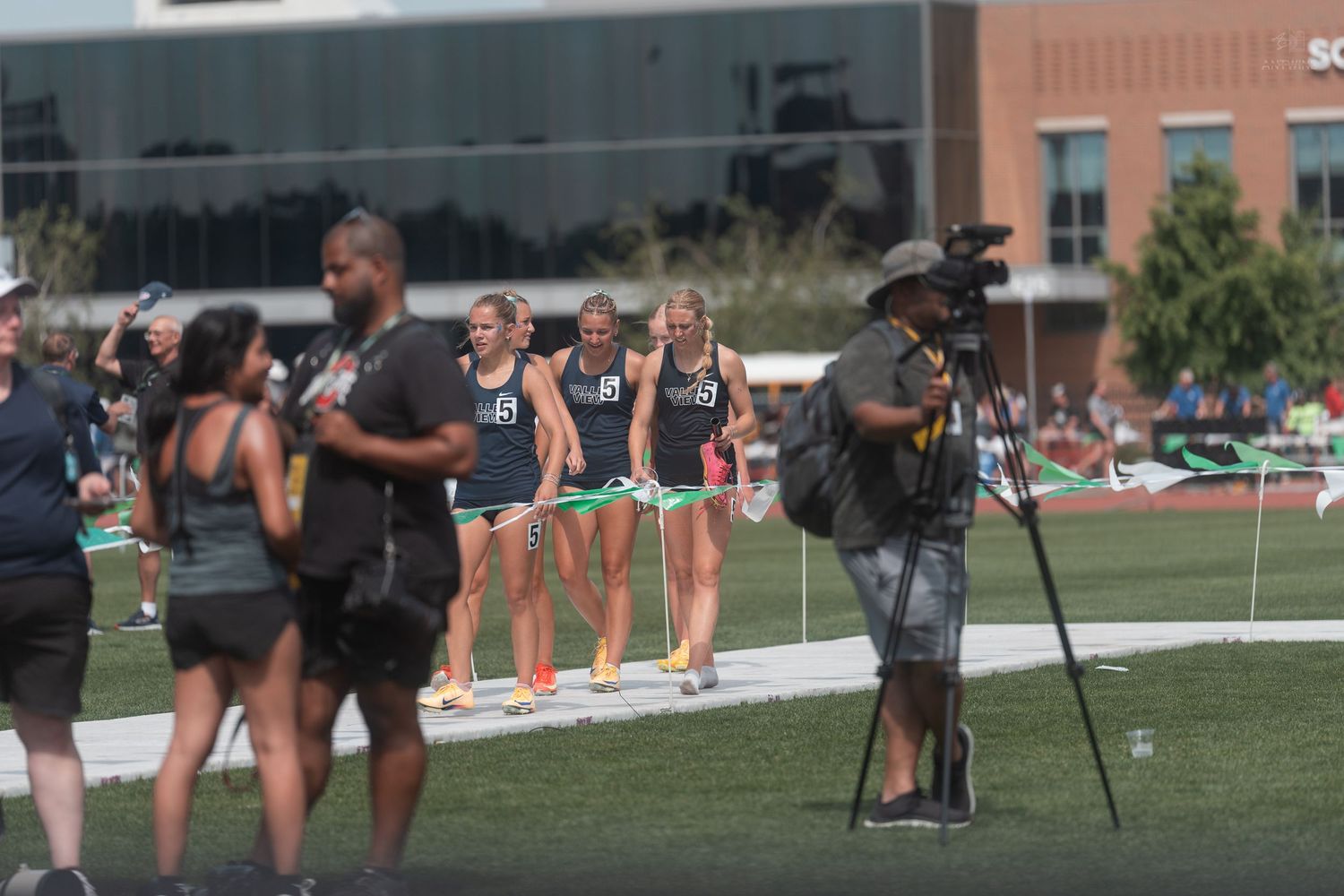 Female track athletes in dark uniforms being filmed by a videographer at an outdoor track meet.