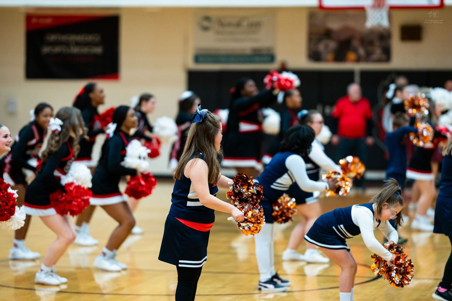 Cheerleaders with red and gold pom-poms perform on a gymnasium floor during a basketball game event.