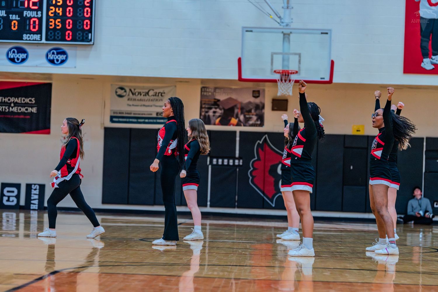 High school cheerleaders in red and black uniforms perform an upbeat routine on a basketball court sideline.