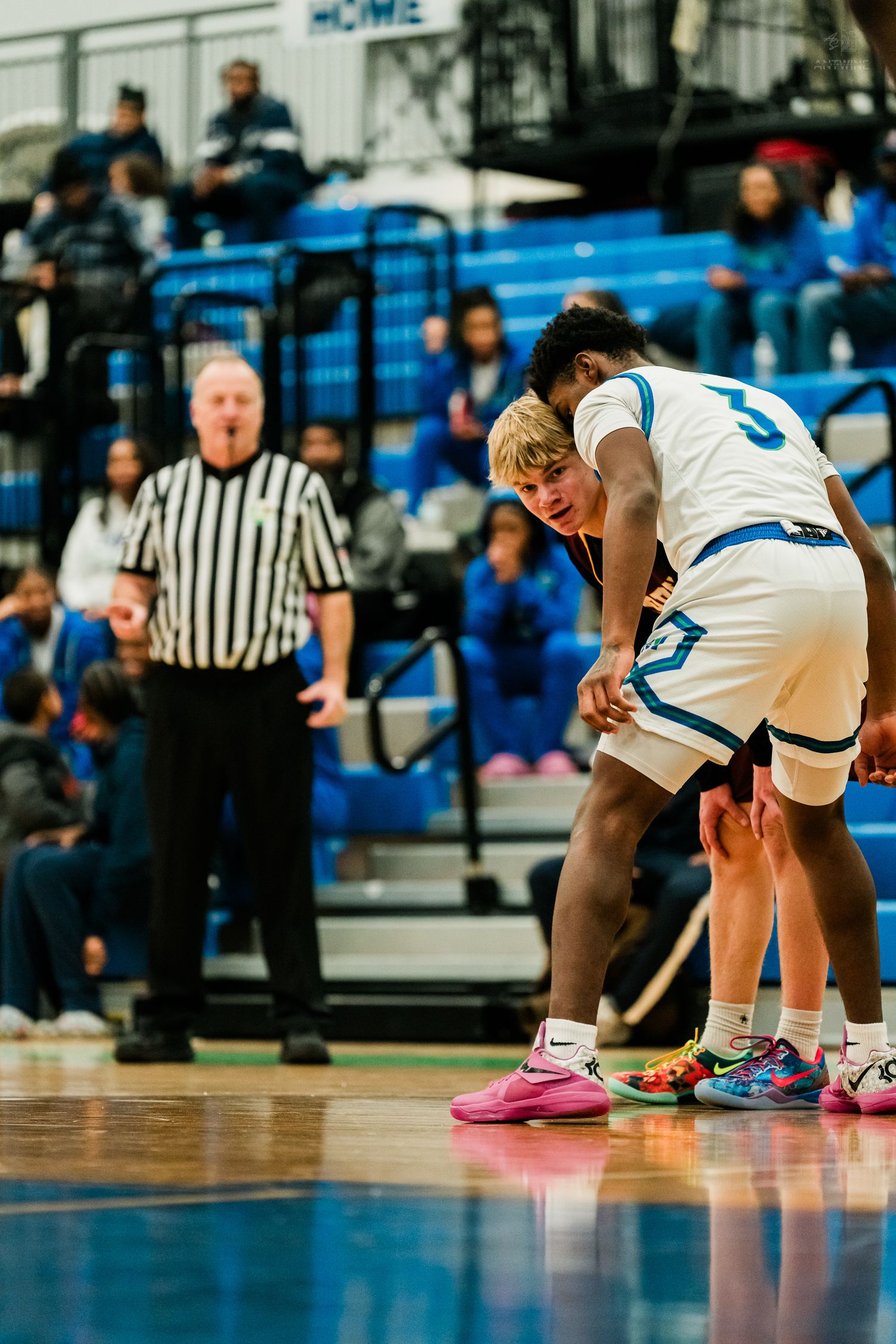 Two basketball players in white uniforms strategize together while referee watches during high school game.
