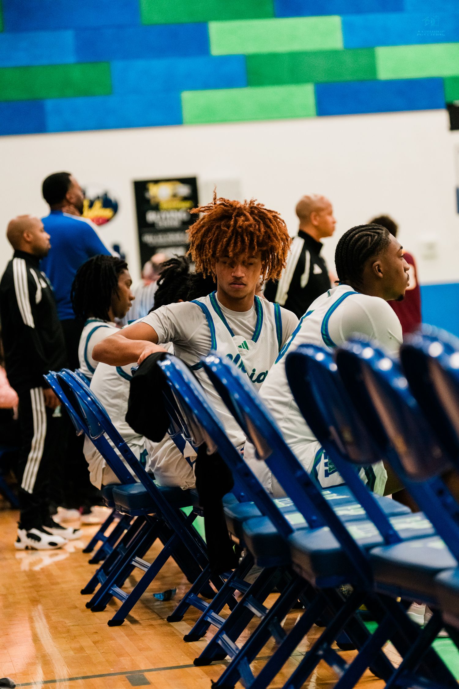 Basketball player with red dreadlocks stands near blue chairs in gym.