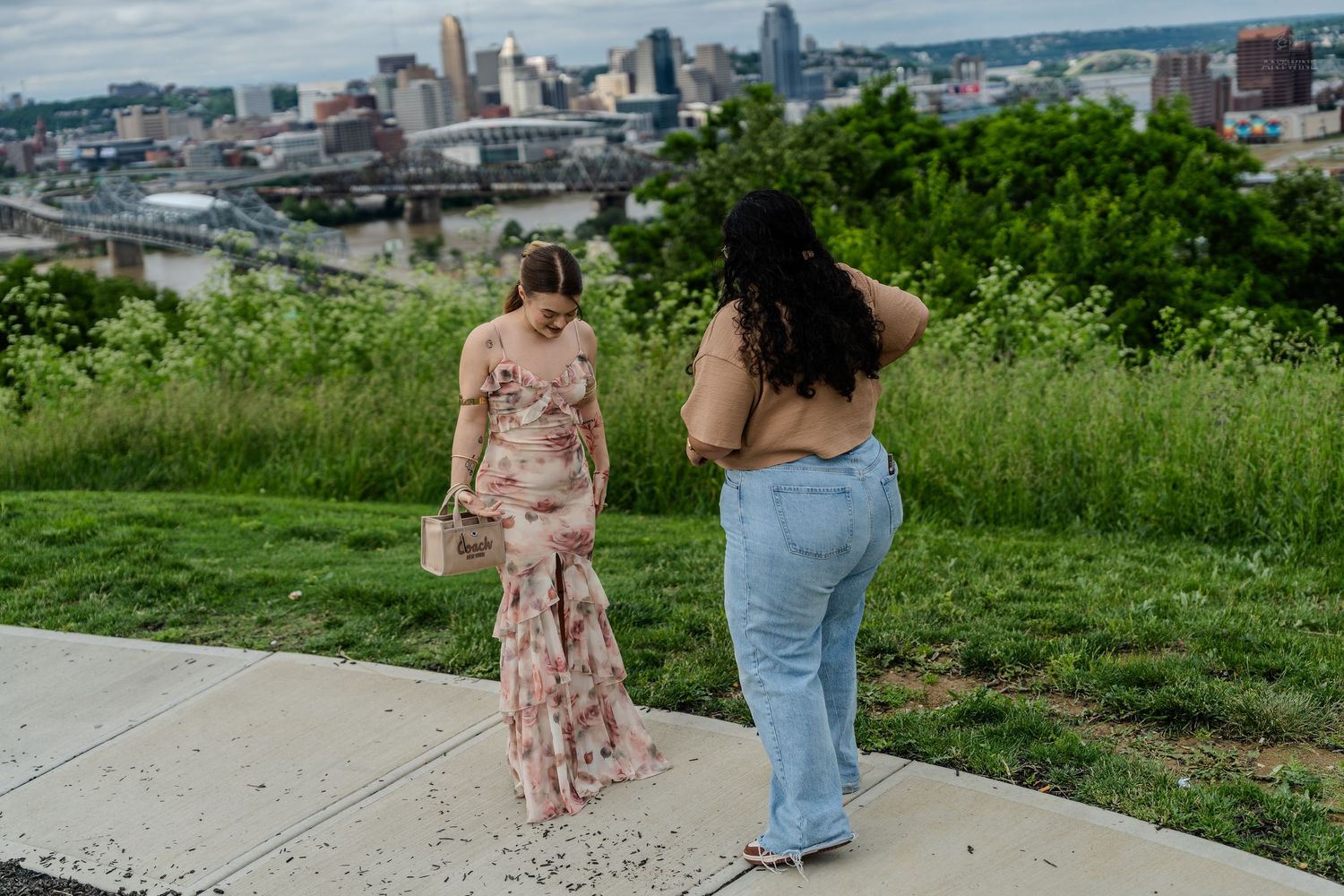 Two women walk along a scenic overlook path with a city skyline and river visible behind them.
