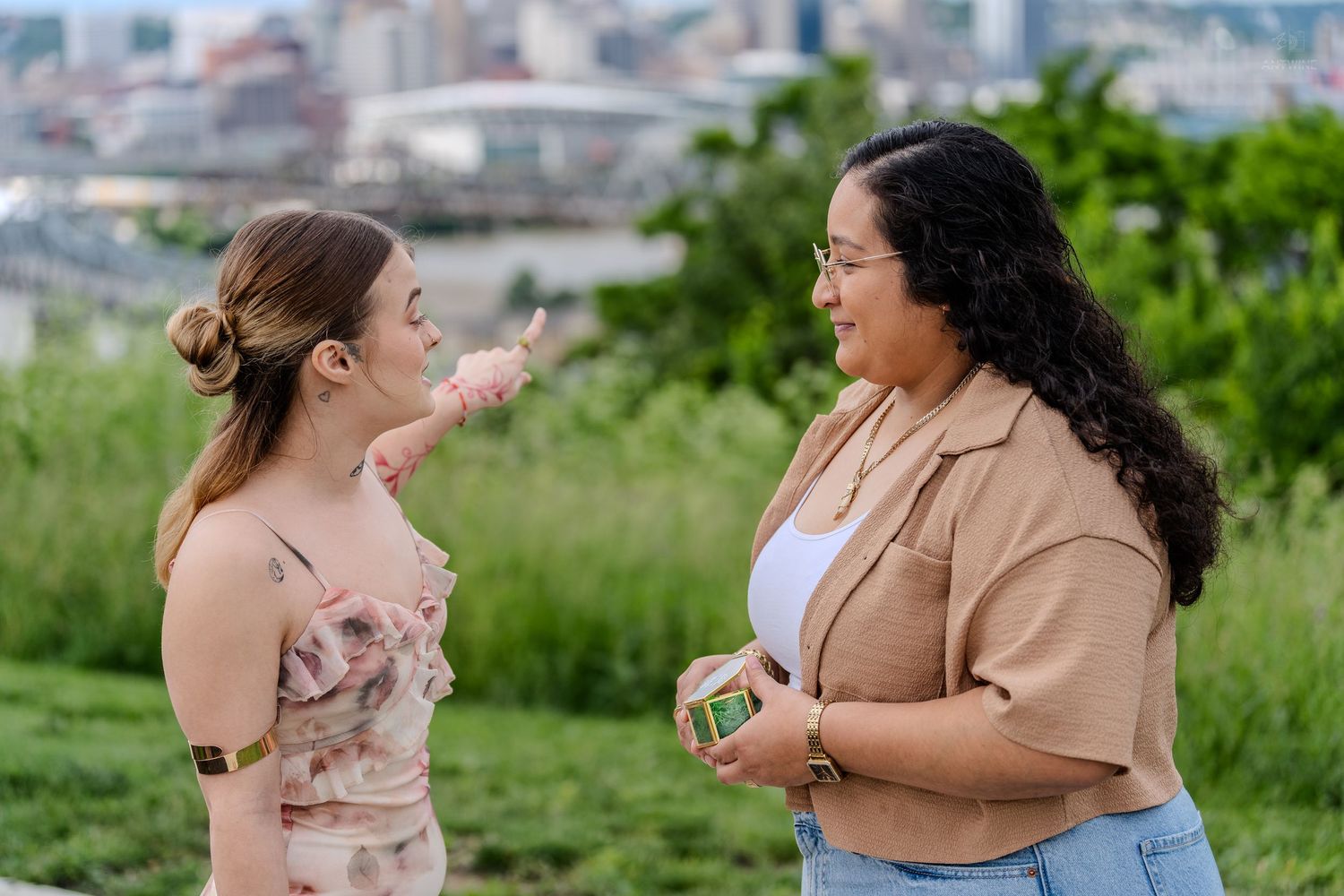 Two women talking and gesturing outdoors with a blurred city skyline backdrop.