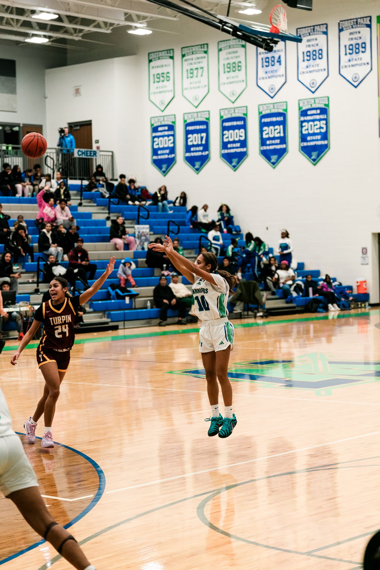 Warriors #10 launches jump shot over Trojan #24 defender with championship banners visible in background.