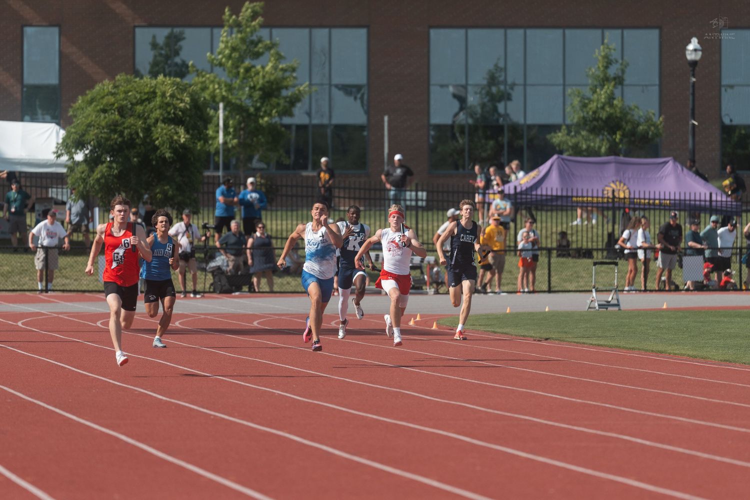 High school athletes compete in a track race on a red running track during an outdoor meet.
