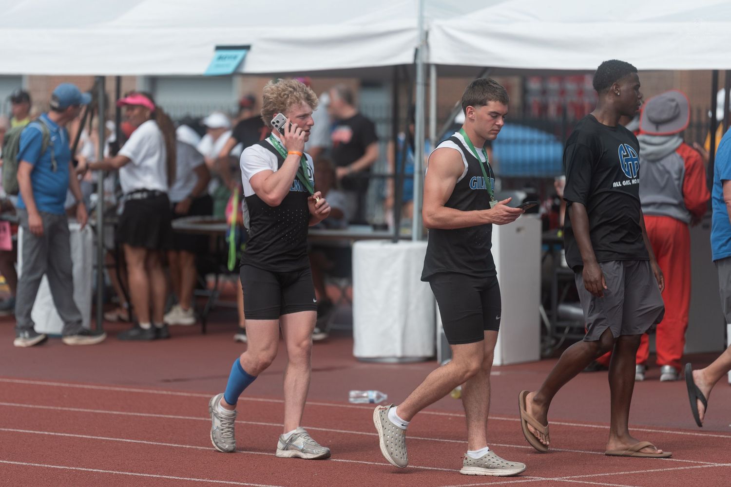 Two male athletes in black and green track uniforms walk along a red track at an outdoor competition.