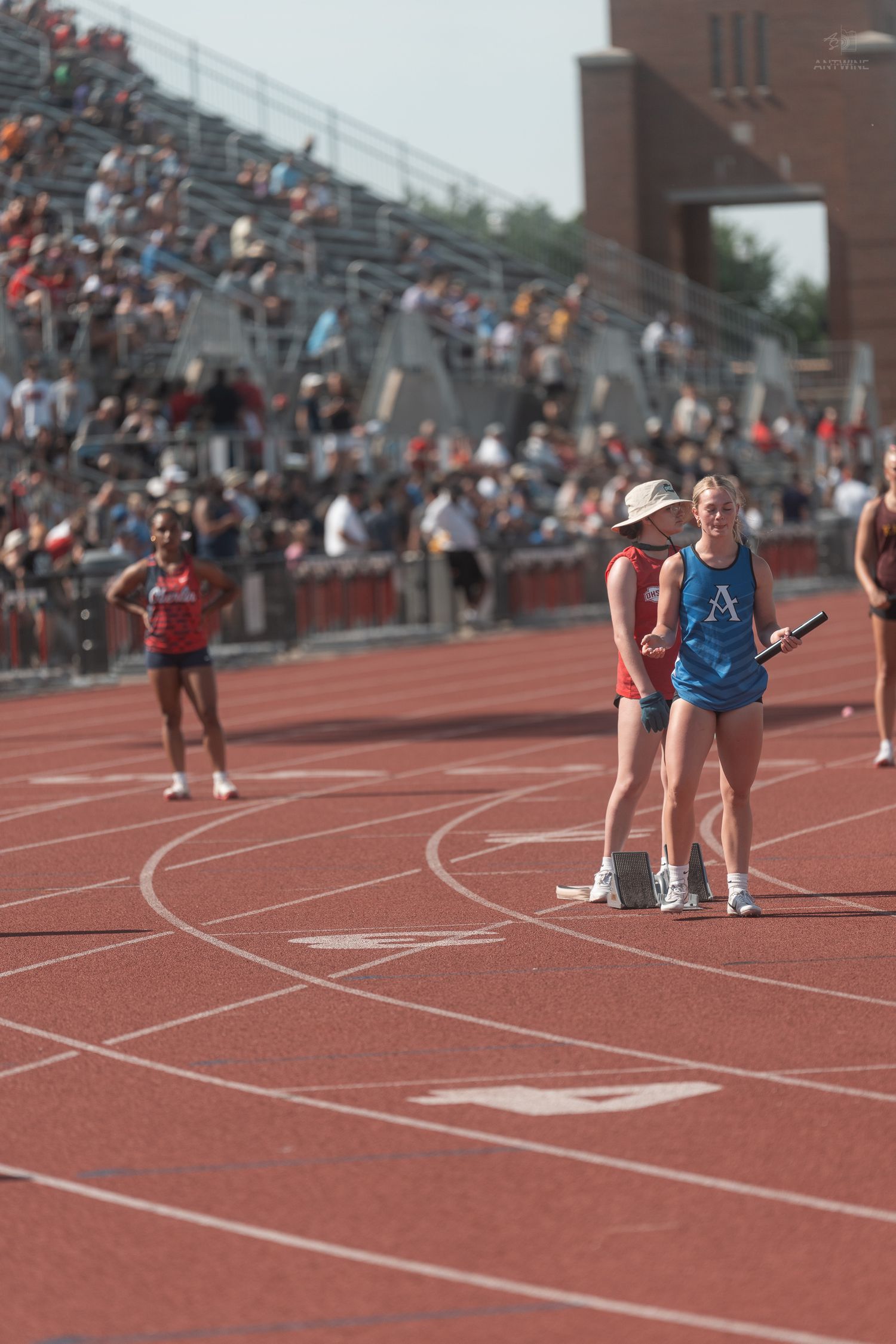 Two athletes stand on a red track holding a relay baton during an outdoor track and field meet.