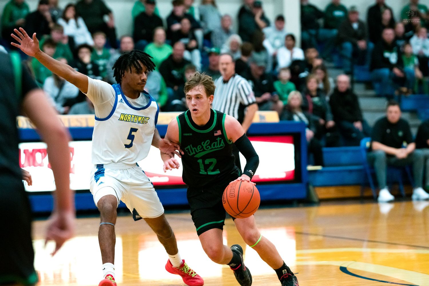 Green-uniformed player drives past a blue-uniformed defender during a high school basketball game.