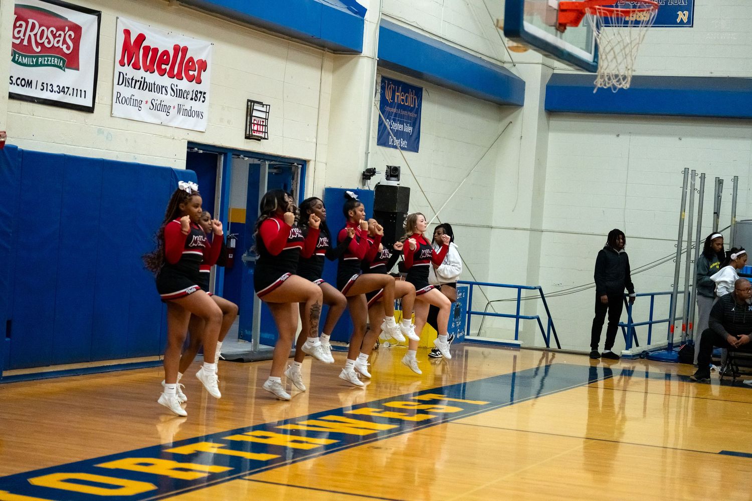High school cheerleaders in red uniforms perform a jumping cheer routine during a basketball game in a gymnasium.